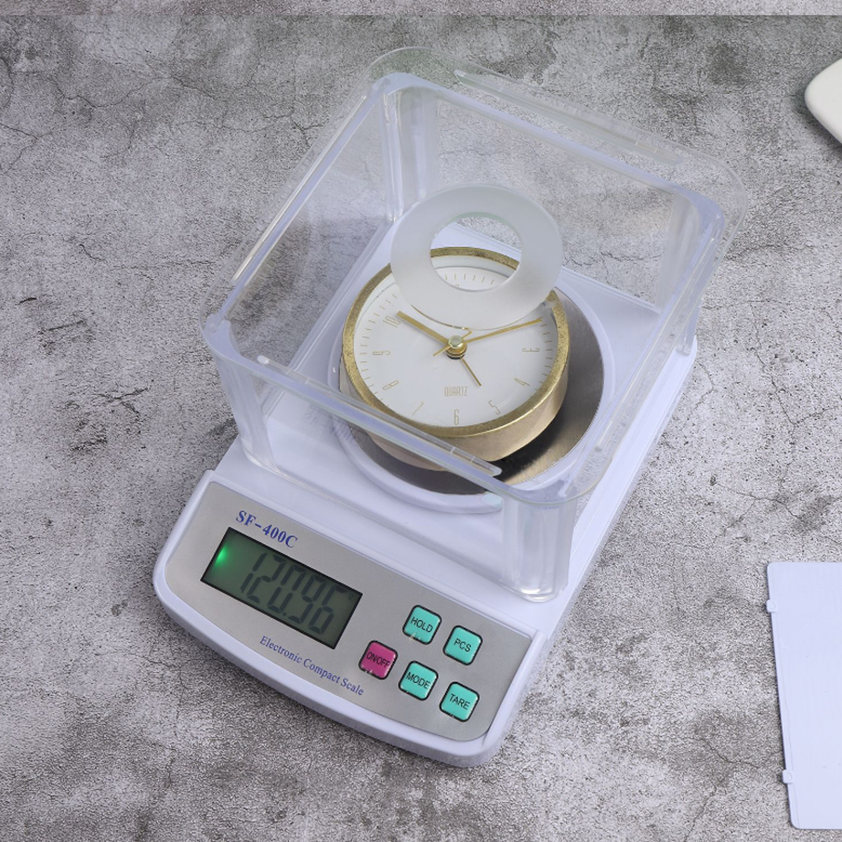 Eine Digitalwaage mit einer Uhr in einer durchsichtigen Box. Die Waage zeigt 120,96 auf ihrem grün beleuchteten Display an.