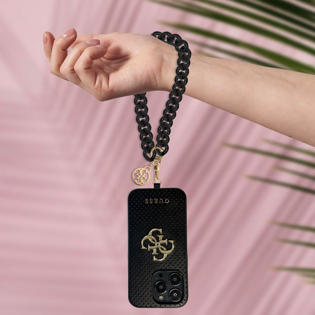 Una mano tiene in mano un telefono con una custodia Guess nera, una catena nera e uno sfondo rosa.