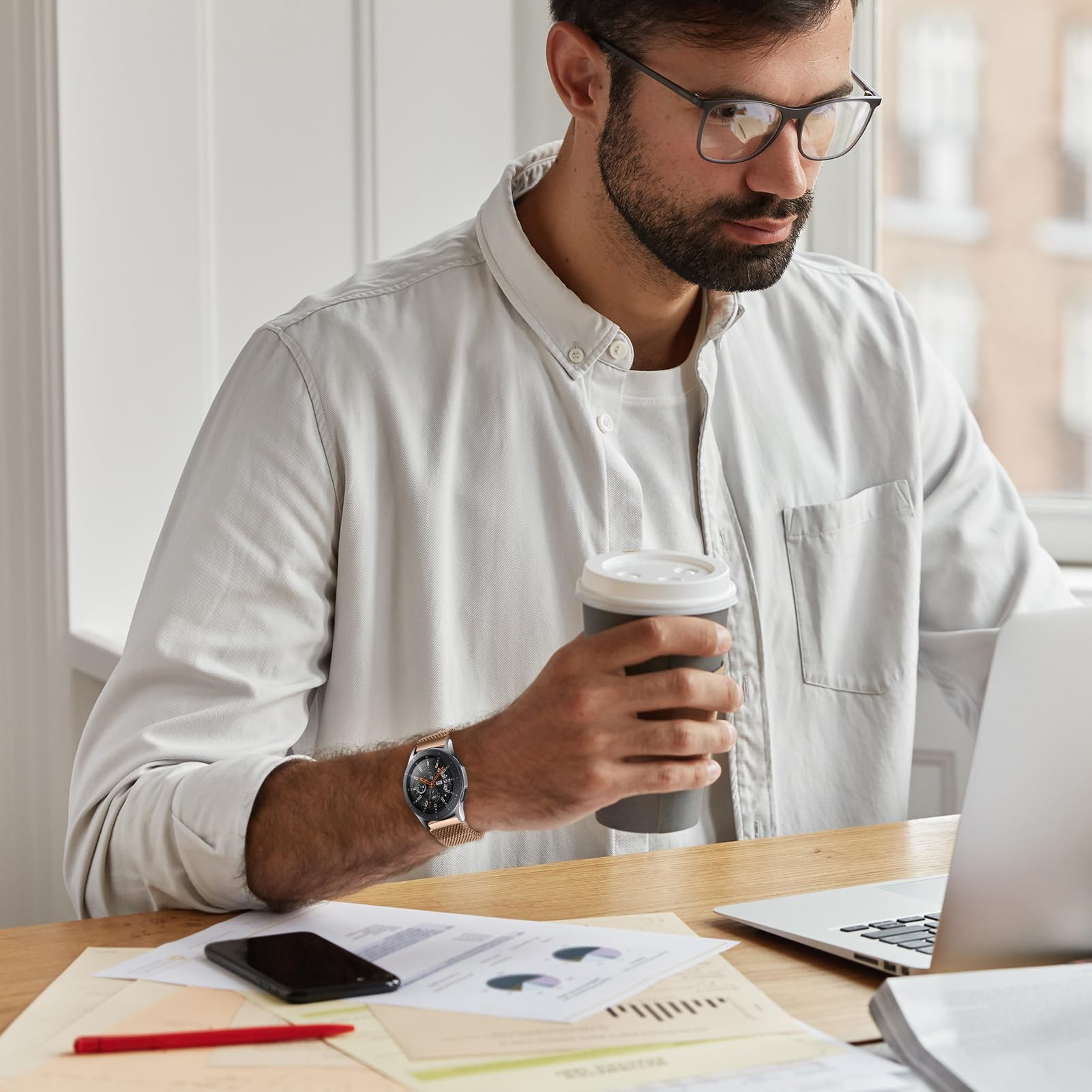 Mann mit Brille schaut auf Laptop, hält Kaffeetasse, Uhr am Handgelenk, Dokumente auf dem Schreibtisch.