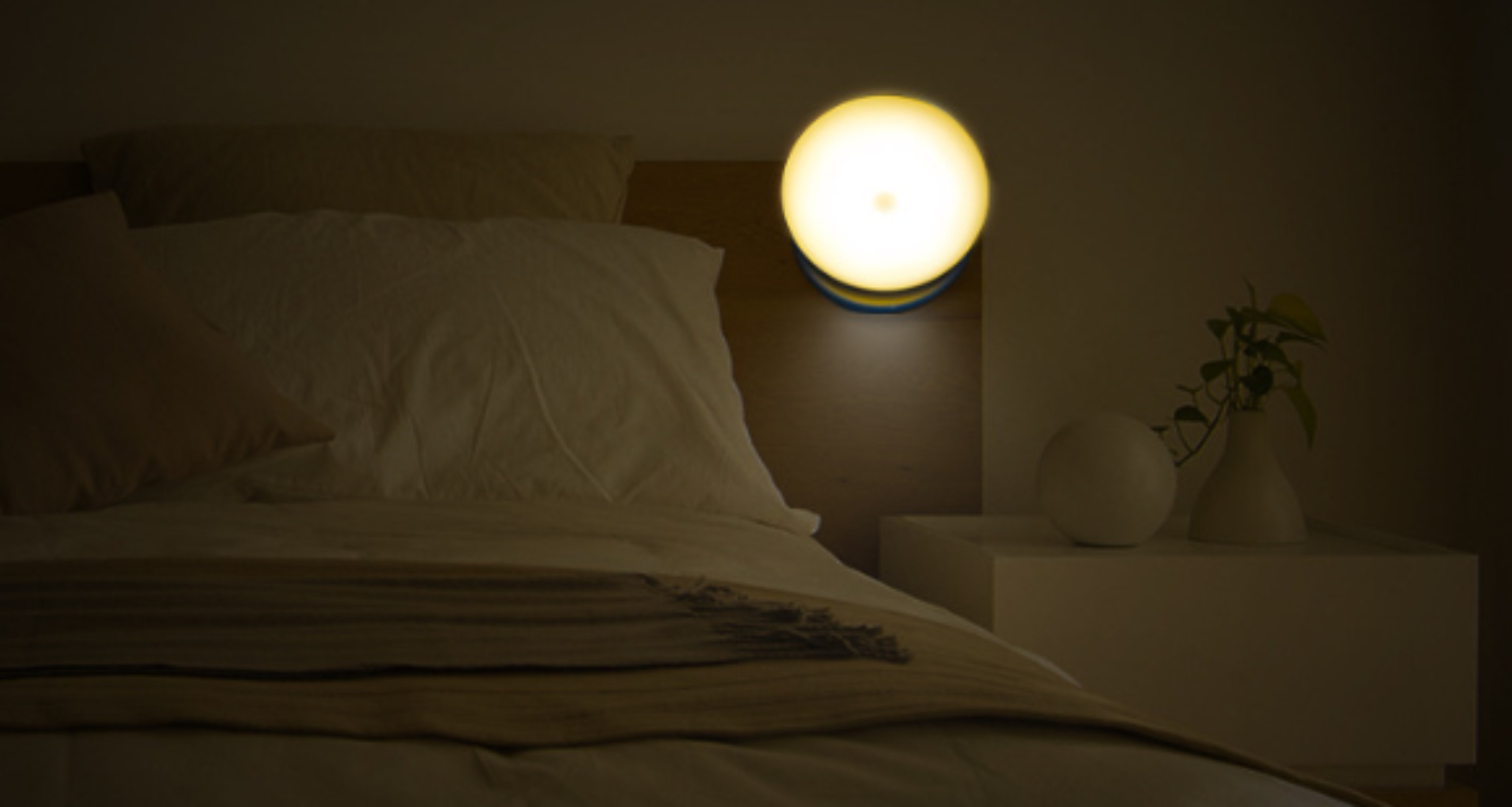 Schlafzimmer bei Nacht mit rundem Nachtlicht an der Wand über dem Bett, mit Pflanze und Deko.