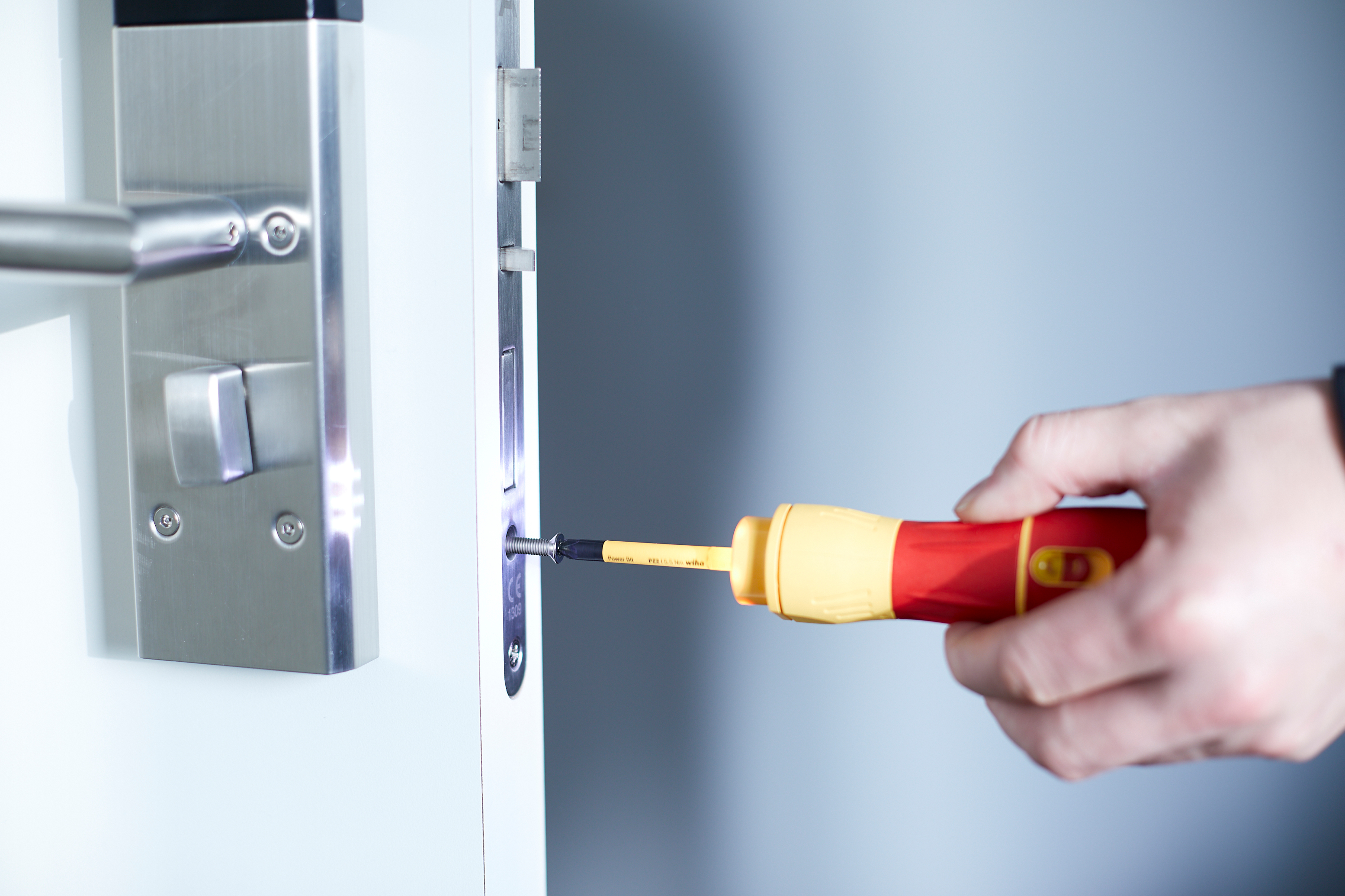 Osoba używająca śrubokręta do zabezpieczenia zamka do drzwi, białe drzwi i ościeżnica w tle.