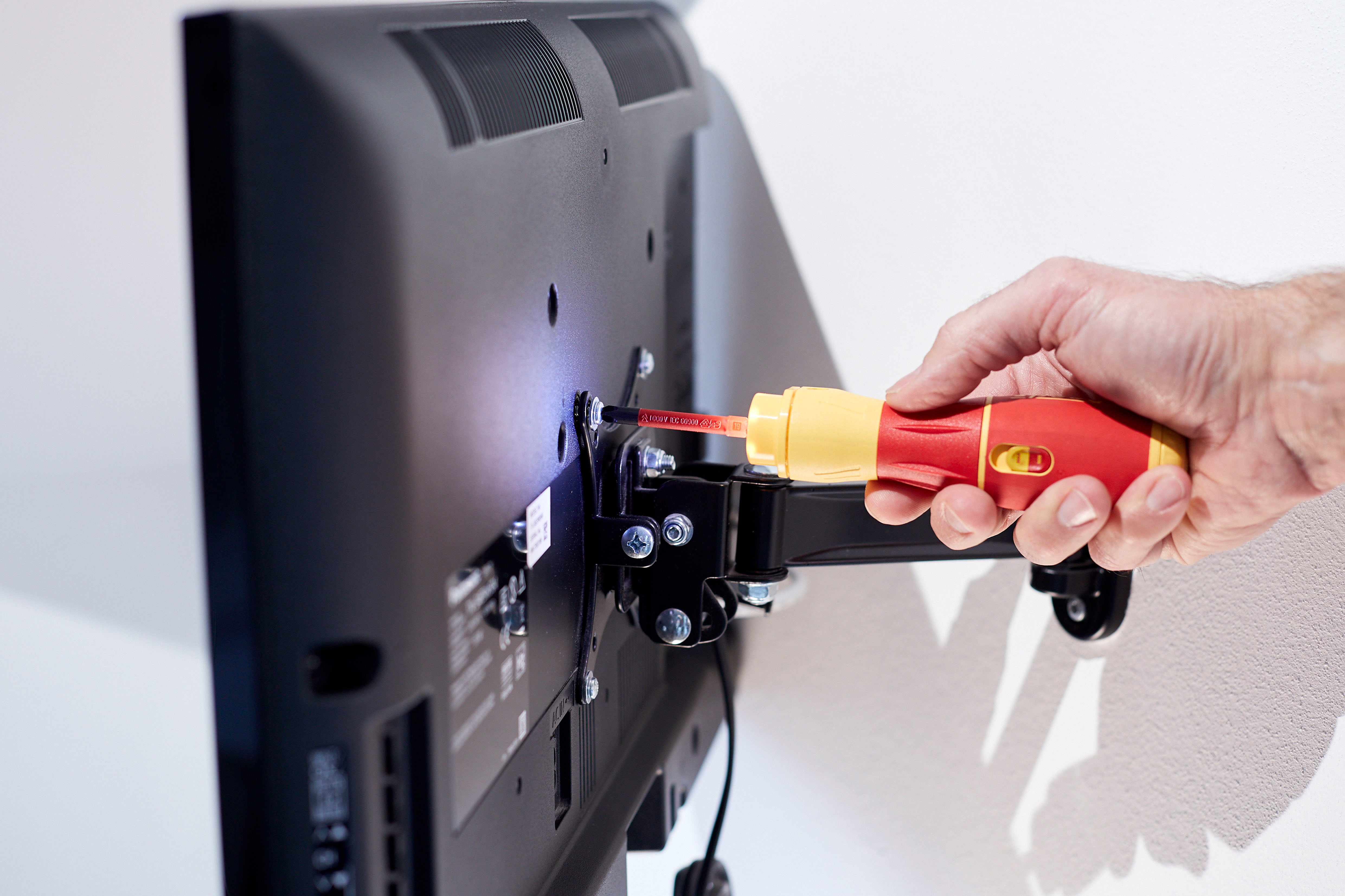 Osoba używająca śrubokrętu do montażu telewizora na uchwycie ściennym, na białej ścianie.