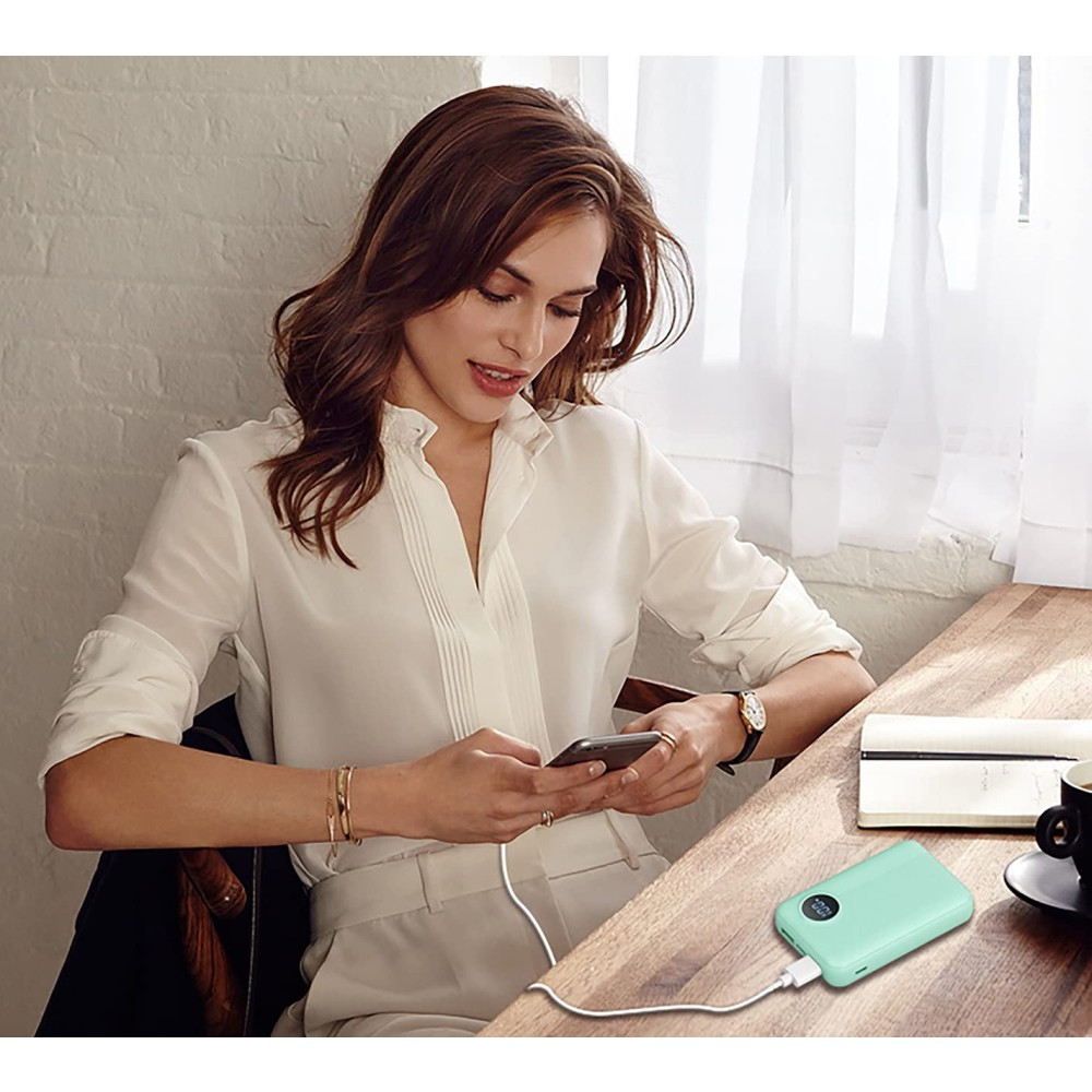 Donna usa telefono, carica da power bank verde menta. Seduta a tavolo con tazza di caffè.