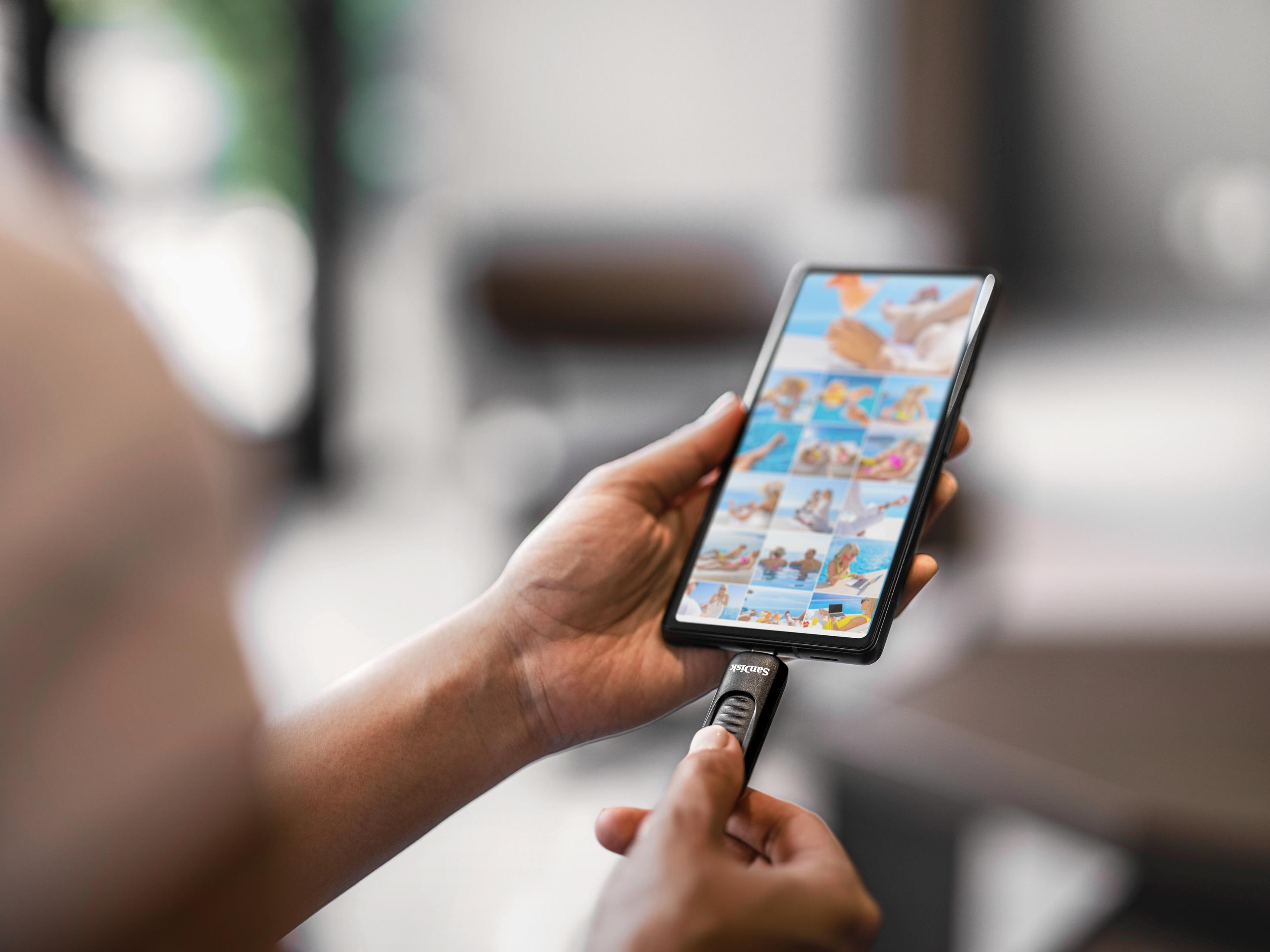 Osoba trzymająca telefon z napędem USB. Telefon wyświetla zdjęcia. Napęd USB jest czarny i podłączony.