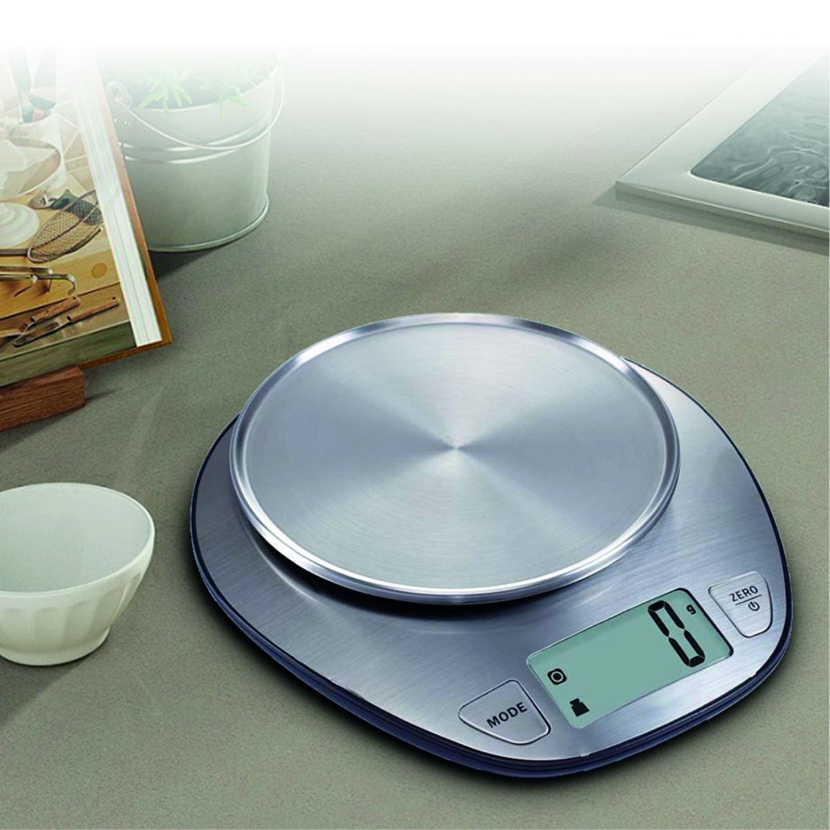 Balanza digital de cocina plateada sobre una superficie gris. Muestra '0' en su pantalla. Se ven otros objetos.