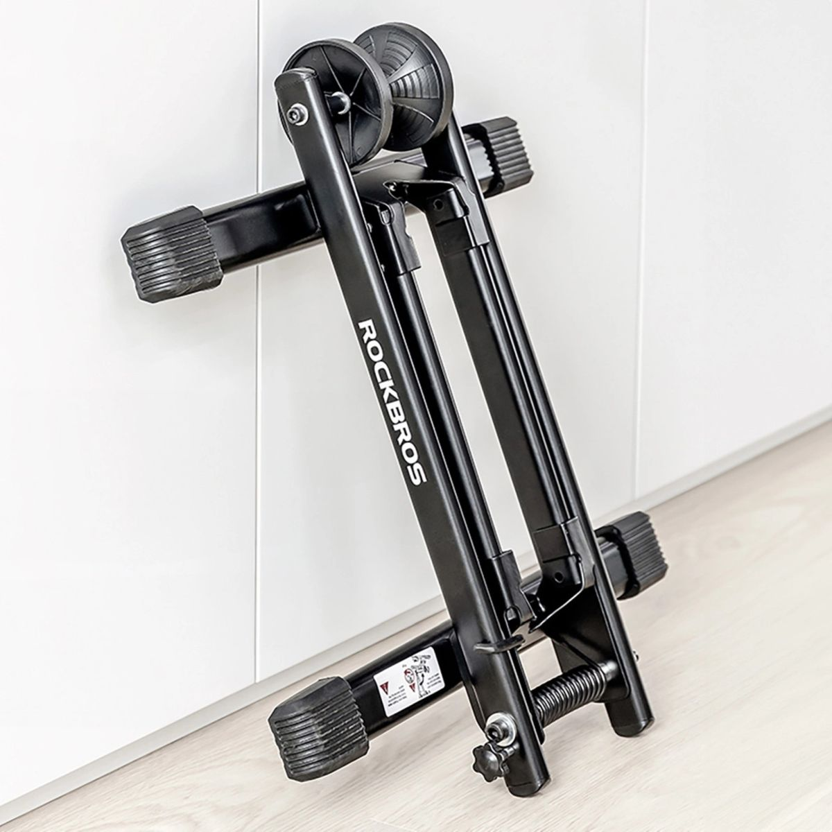 Ein schwarzer Fahrradtrainer-Ständer lehnt an einer weißen Wand. Der Markenname "ROCKBROS" ist sichtbar.