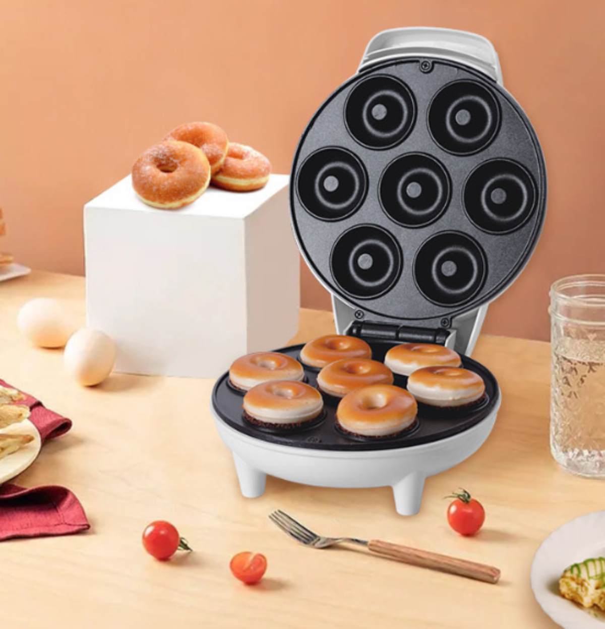 Weiße Donut-Maschine mit Donuts innen und drei daneben, mit Tomaten, Gabel und Wasser.