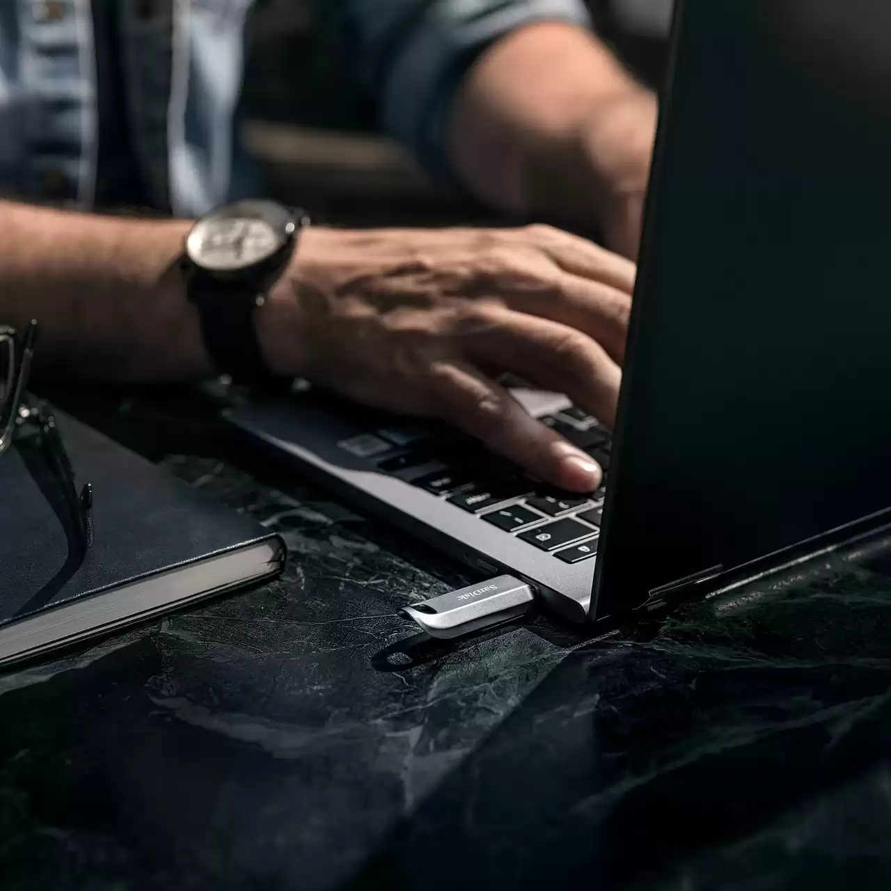 Osoba korzysta z laptopa z podłączonym pendrivem USB Sandisk. Notatnik i okulary.