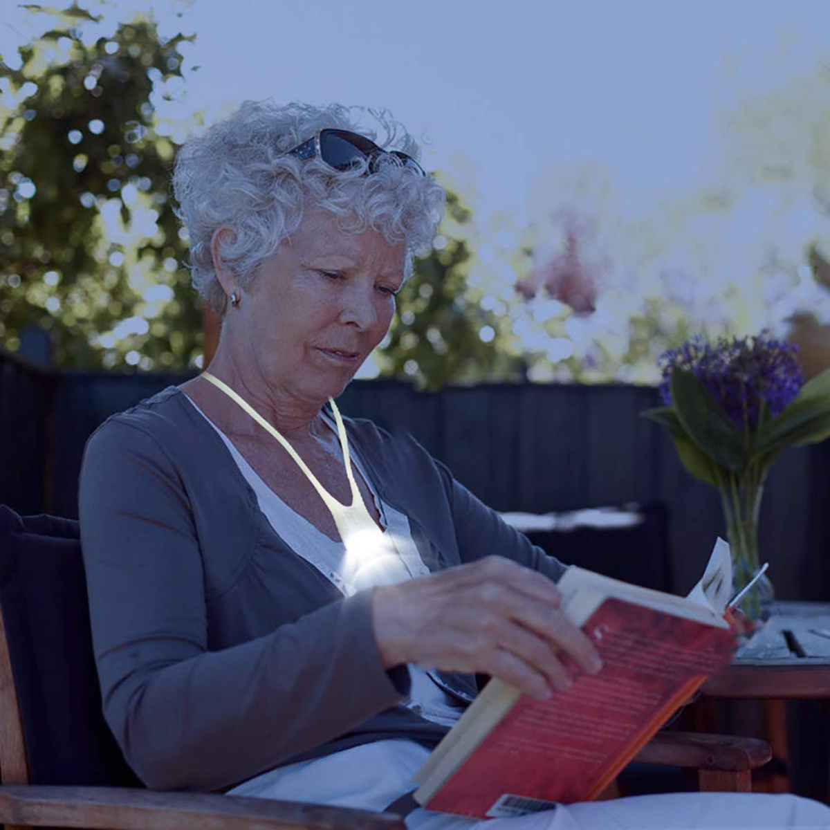 Una mujer mayor leyendo un libro en un jardín de noche, iluminada por una luz que lleva en el cuello.