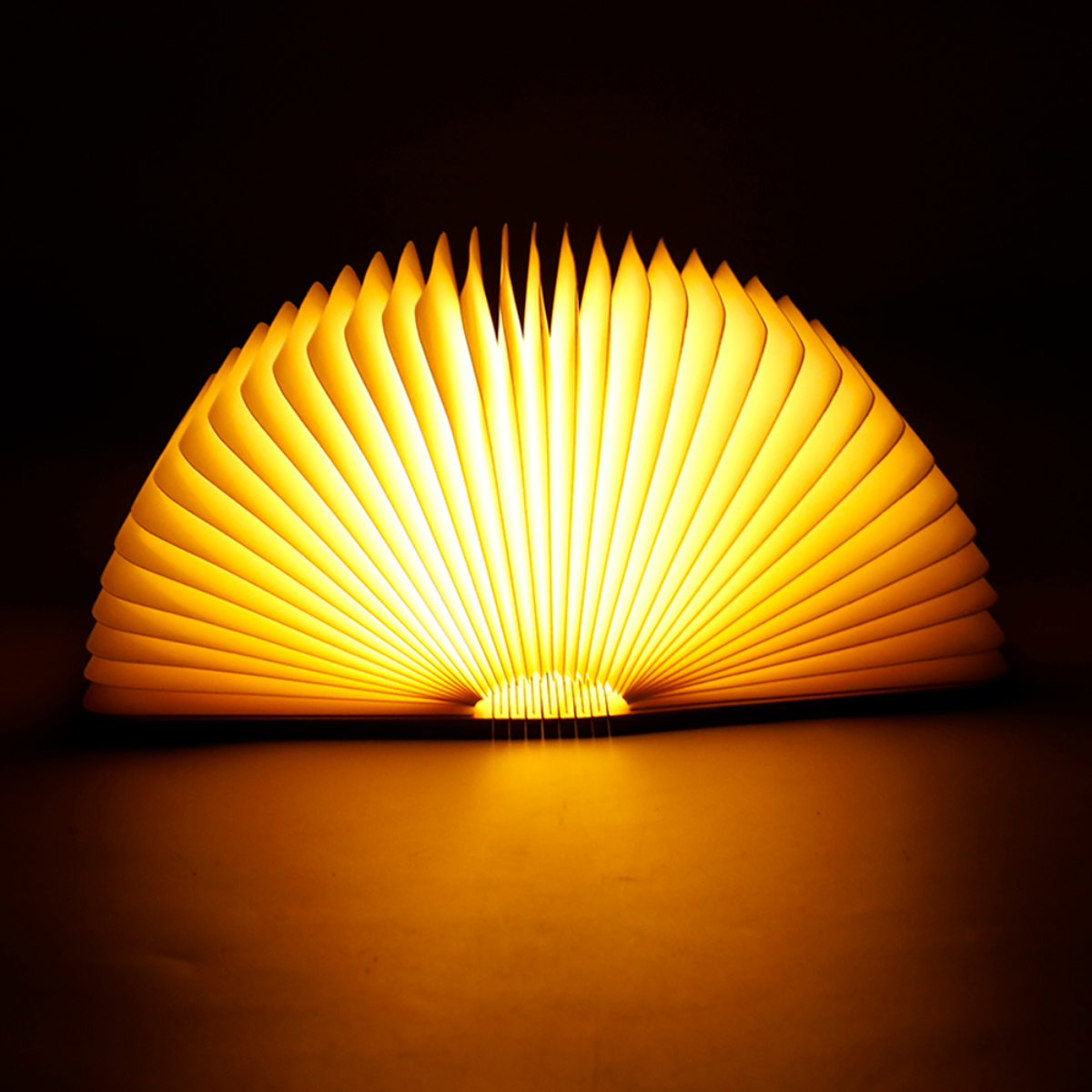 Lámpara con forma de libro abierta, emitiendo una luz cálida y amarilla. La luz brilla sobre una superficie.