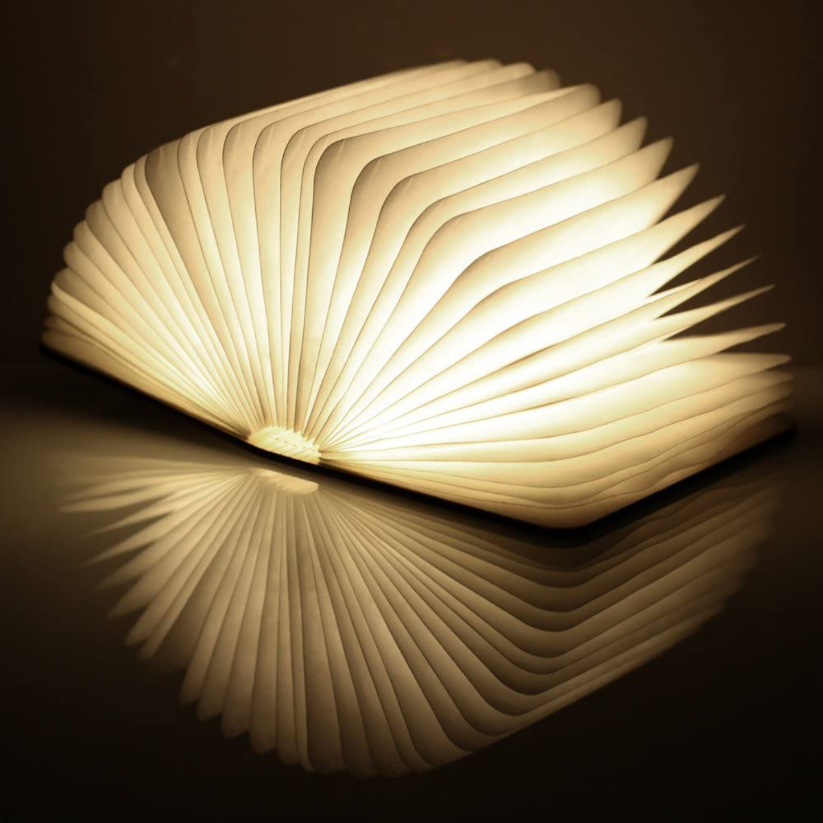 Lámpara con forma de libro, abierta, con brillo cálido y reflejo. La lámpara está sobre una superficie oscura.