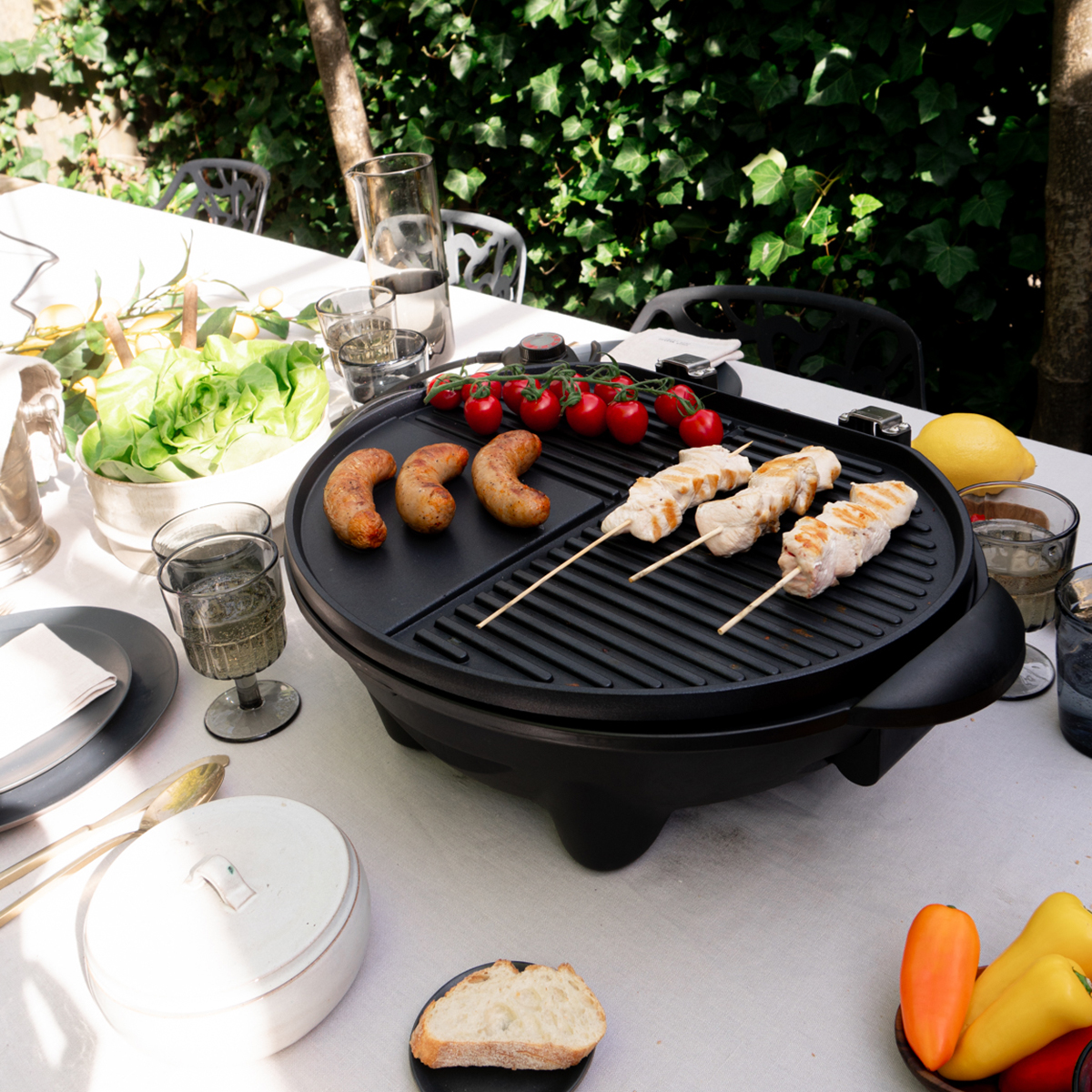 An electric grill on a table with cooked food, vegetables, and glasses of water.