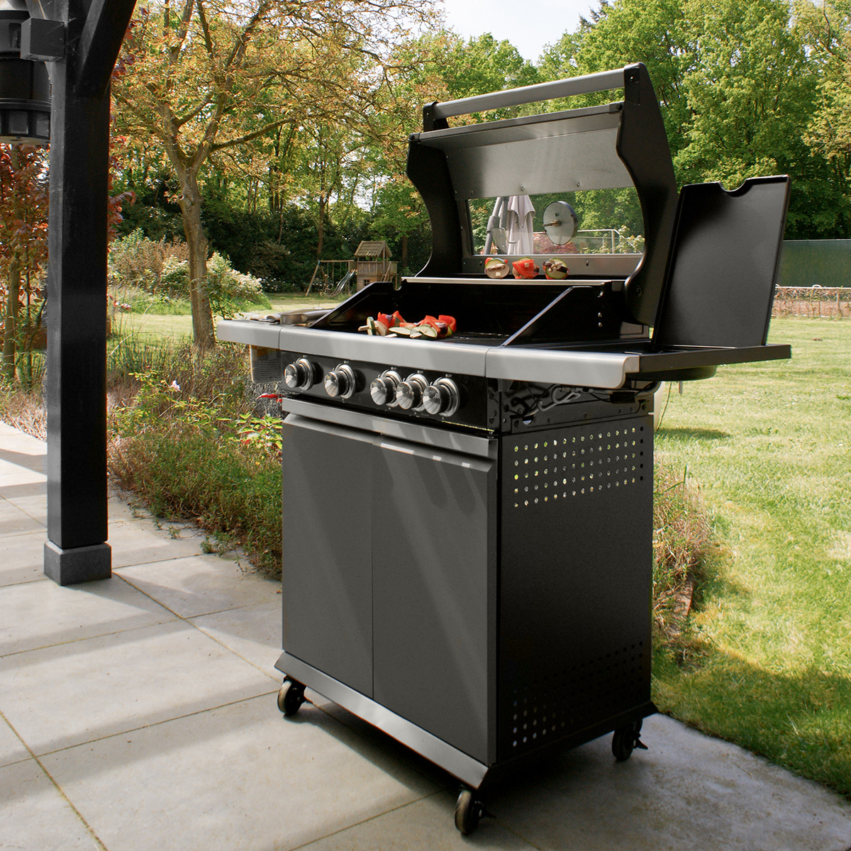 A black grill set up in a garden with food and utensils on the side.