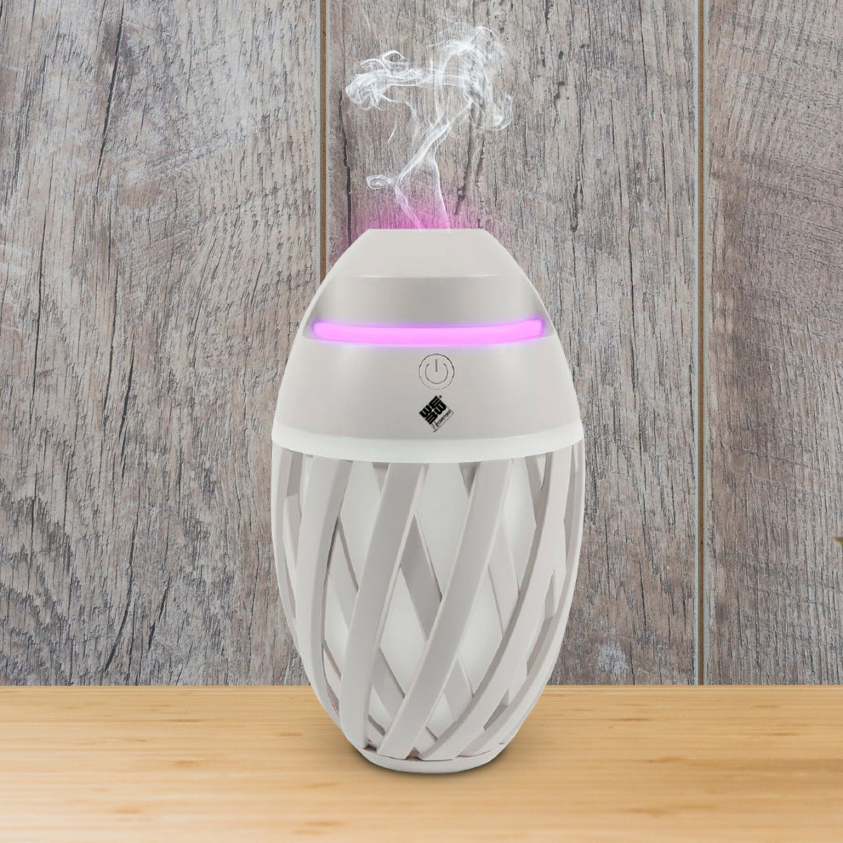Difusor de aroma blanco con luz rosa y humo, sobre superficie de madera. Fondo con pared de madera.