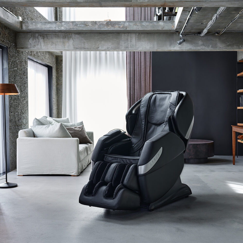 A black massage chair in a modern room. It's in front of a white sofa and near a floor lamp.