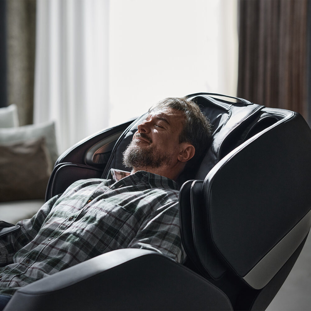 A man relaxes in a black massage chair. He's wearing a plaid shirt and has his eyes closed.