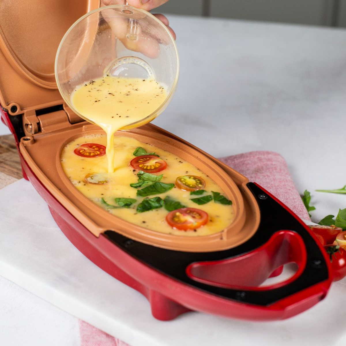 Una mano vierte mezcla de huevo en una omeletera roja abierta con tomates.