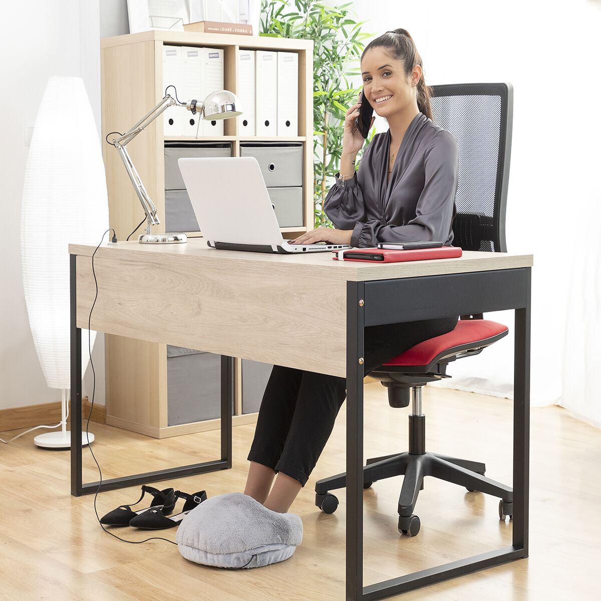 Una mujer está sentada en un escritorio hablando por teléfono, con un portátil y un calentador de pies. La habitación es una oficina.