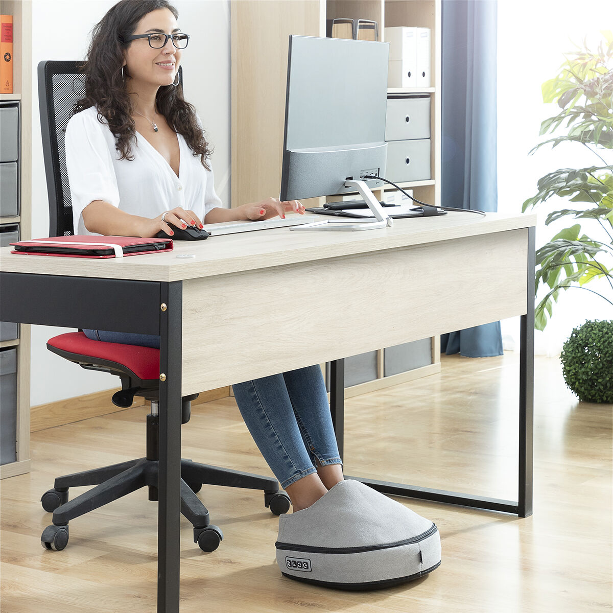 Mujer en escritorio usando computadora, pies en calientapiés. Escritorio y silla visibles.