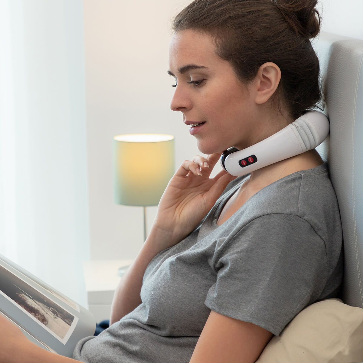 Kobieta w szarej koszulce nosi masażer szyi, czyta książkę. Lampa stoi na stoliku nocnym. Opiera się o wezgłowie.