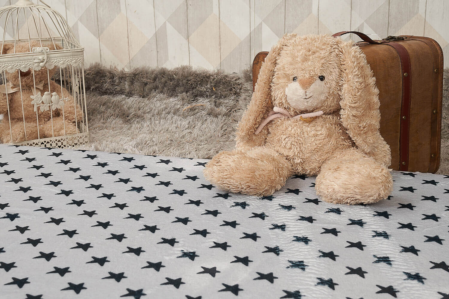 Un conejito de peluche sobre una manta con estrellas, una maleta y una jaula en el fondo.
