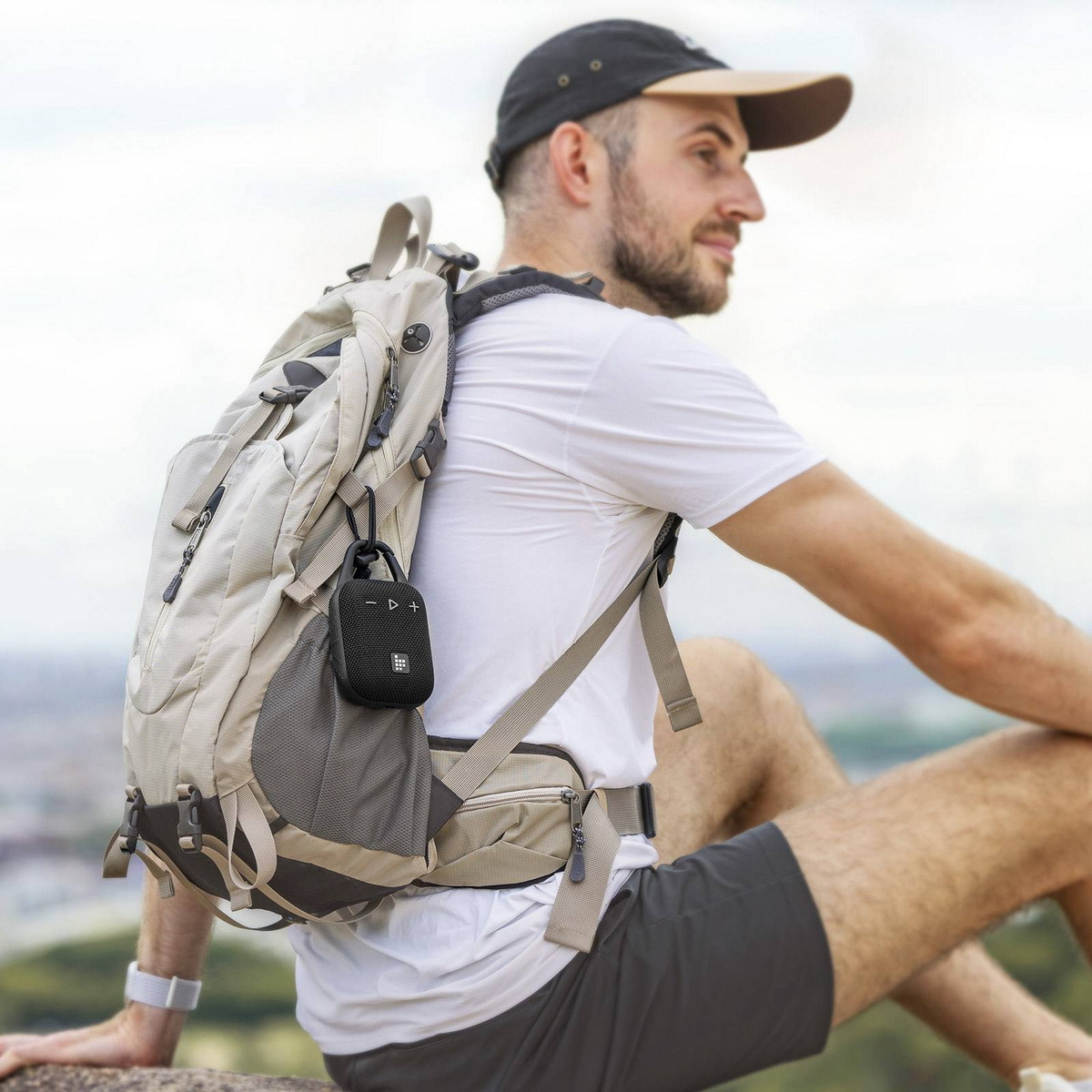 Mann mit Rucksack beim Wandern, Lautsprecher befestigt. Grüne Landschaft im Hintergrund mit blauem Himmel. Der Lautsprecher ist schwarz.