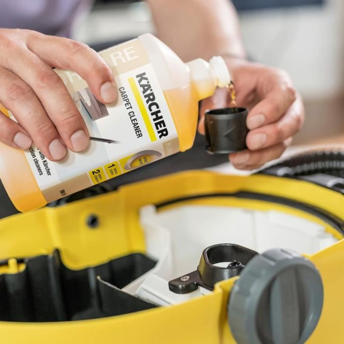 Person gießt Kärcher Teppichreiniger in eine gelbe Maschine. Die Flasche ist mit Kärcher Teppichreiniger beschriftet.