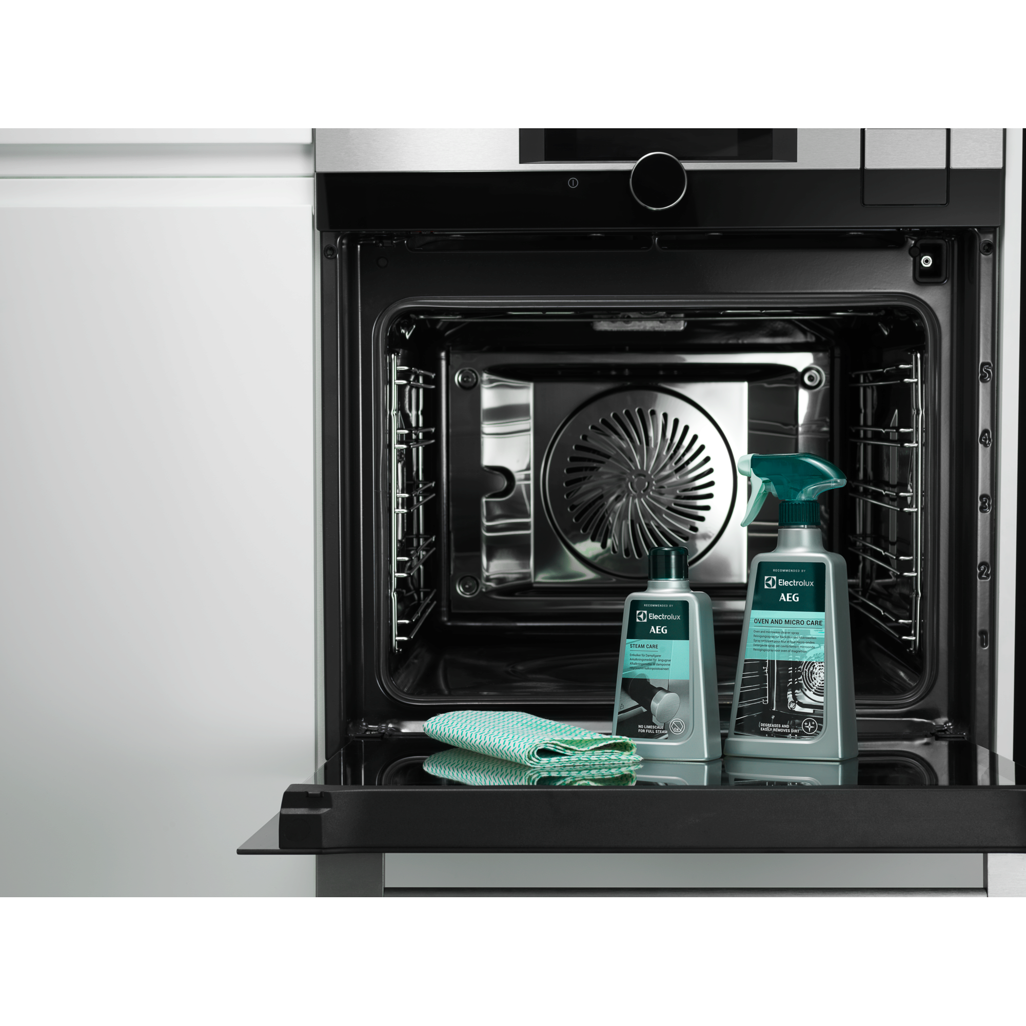 Wnętrze piekarnika ze środkami czyszczącymi i zieloną szmatką. Drzwi otwarte. Szare wykończenie stalowe.
