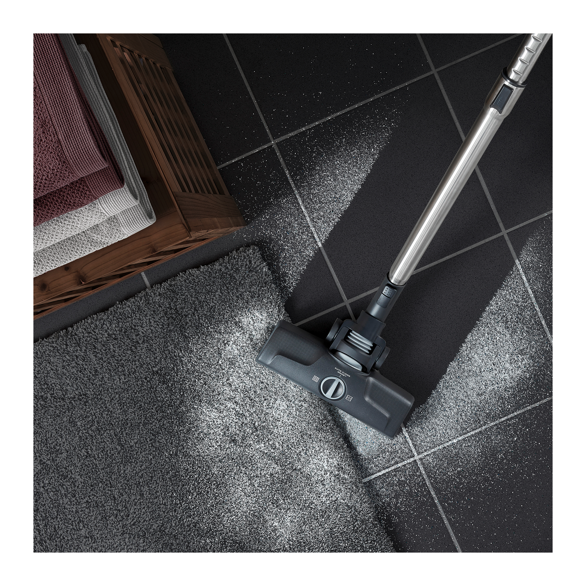 Odkurzacz czyszczący szary dywan na czarnej podłodze z płytek. Ręczniki i kosz w tle.