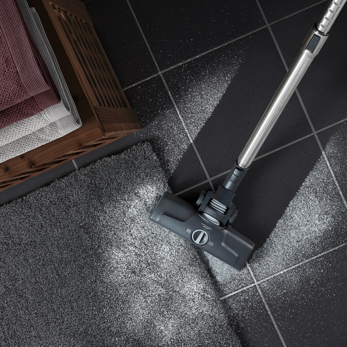 Odkurzacz czyszczący szary dywan na czarnej podłodze z płytek. Ręczniki i kosz w tle.