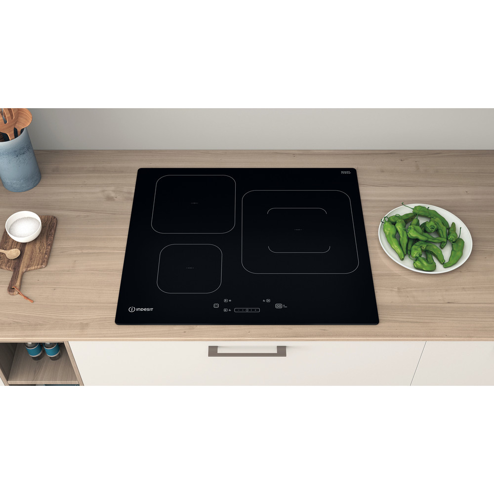 Placa de inducción negra sobre encimera de madera. Pimientos verdes en plato a la derecha.