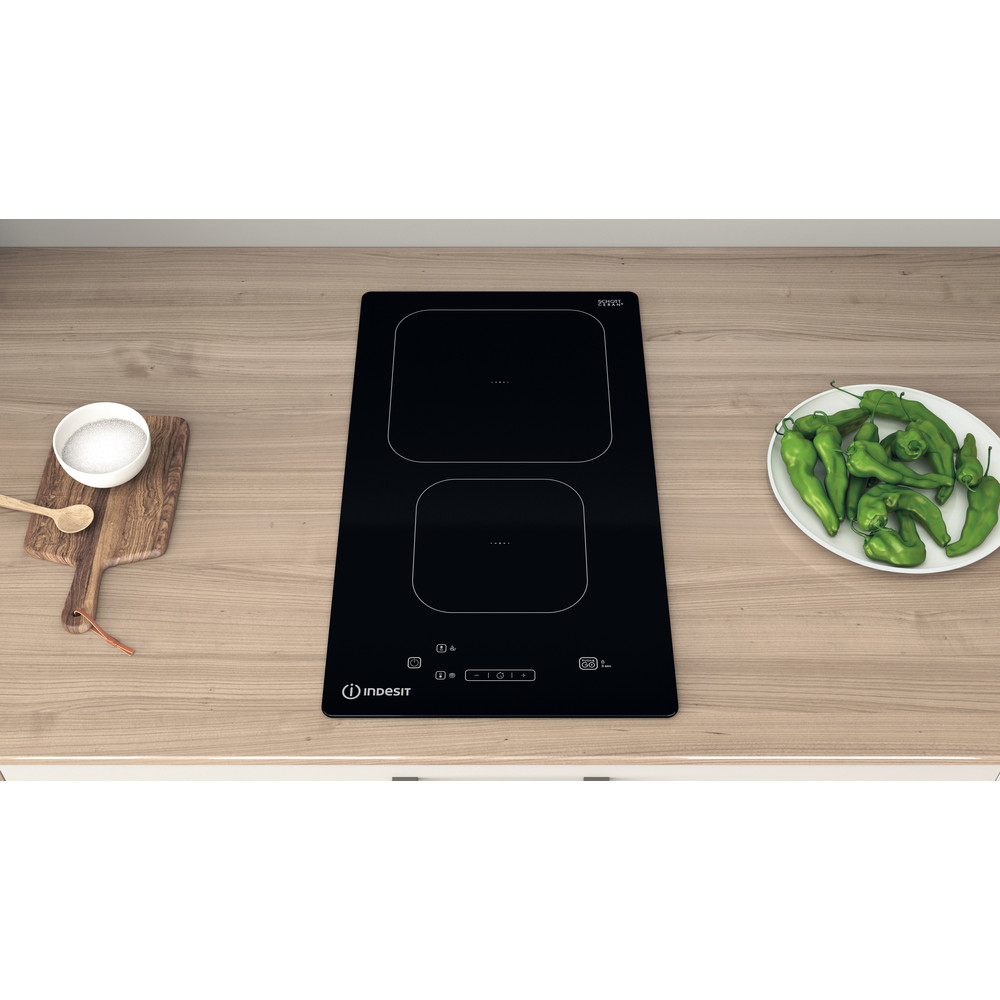 Piano cottura a induzione Indesit nero, tagliere e piatto con peperoni.