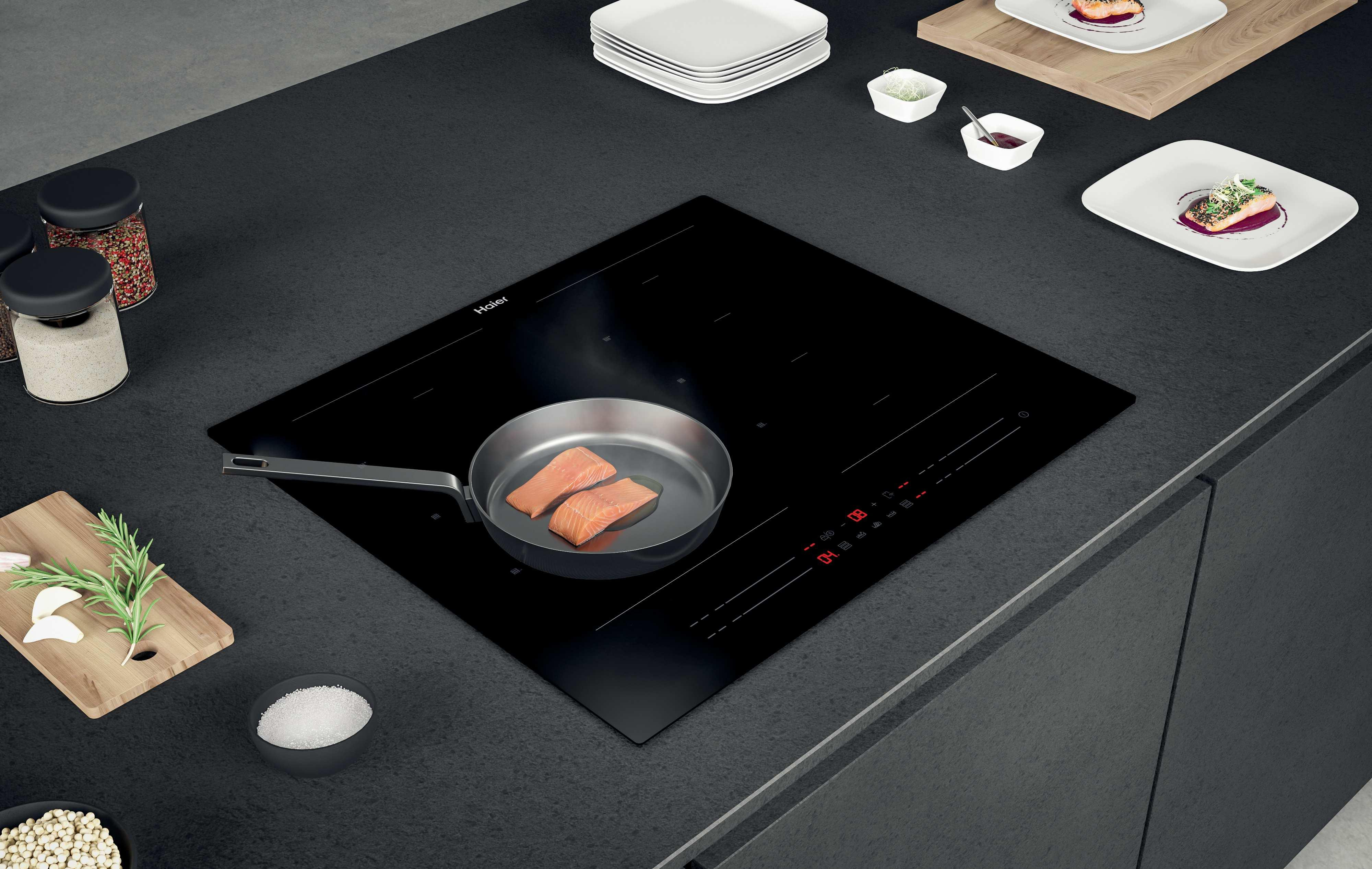 Płyta indukcyjna z patelnią z gotującym się łososiem, talerzami i przyprawami, na czarnym blacie.