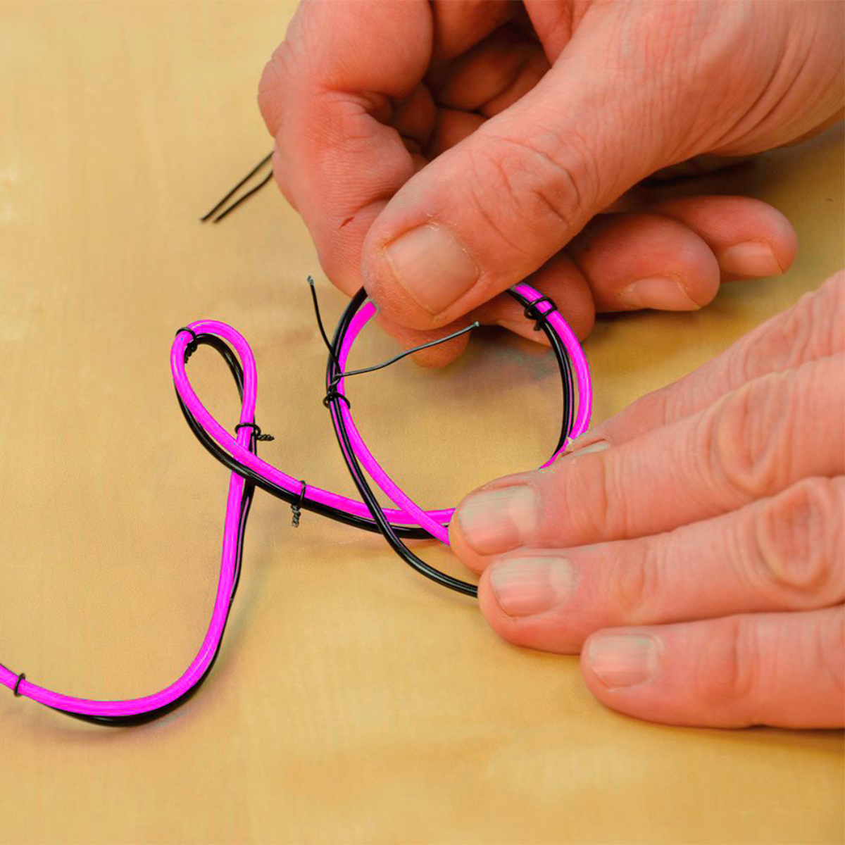 Manos trabajando con un cable de luz de neón rosa, torciéndolo y dándole forma sobre un fondo neutral.