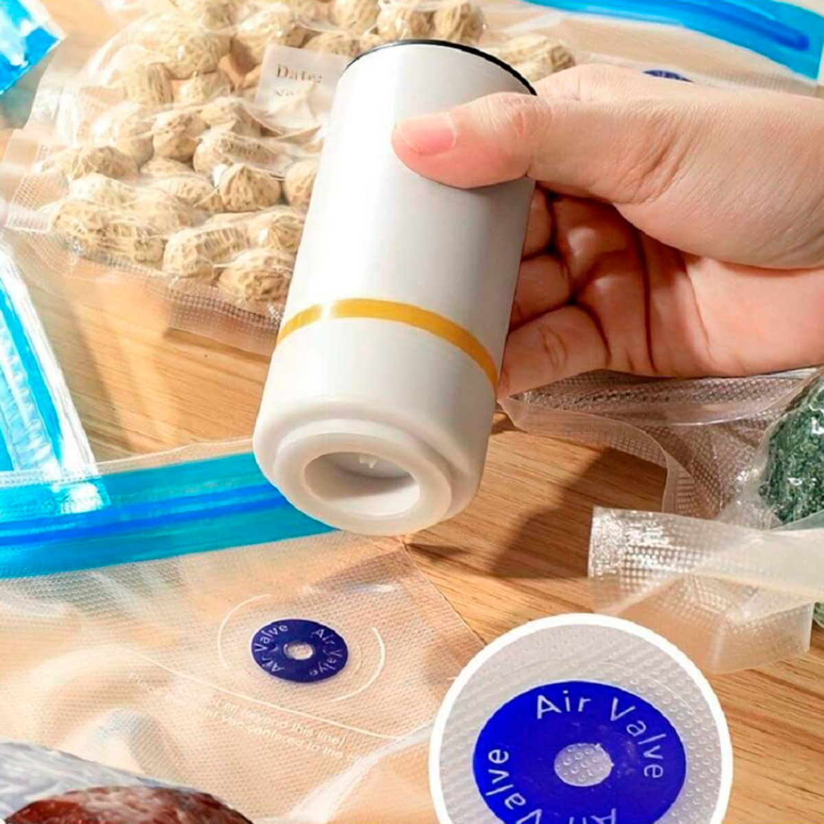 La mano de una persona sosteniendo un dispositivo de vacío portátil blanco cerca de varios alimentos en bolsas selladas.