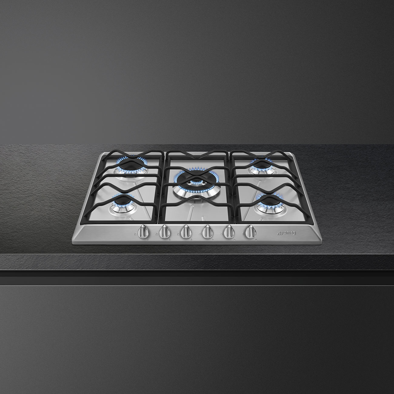 Piano cottura argentato con bruciatori accesi con fiamme blu. Le manopole sono visibili.