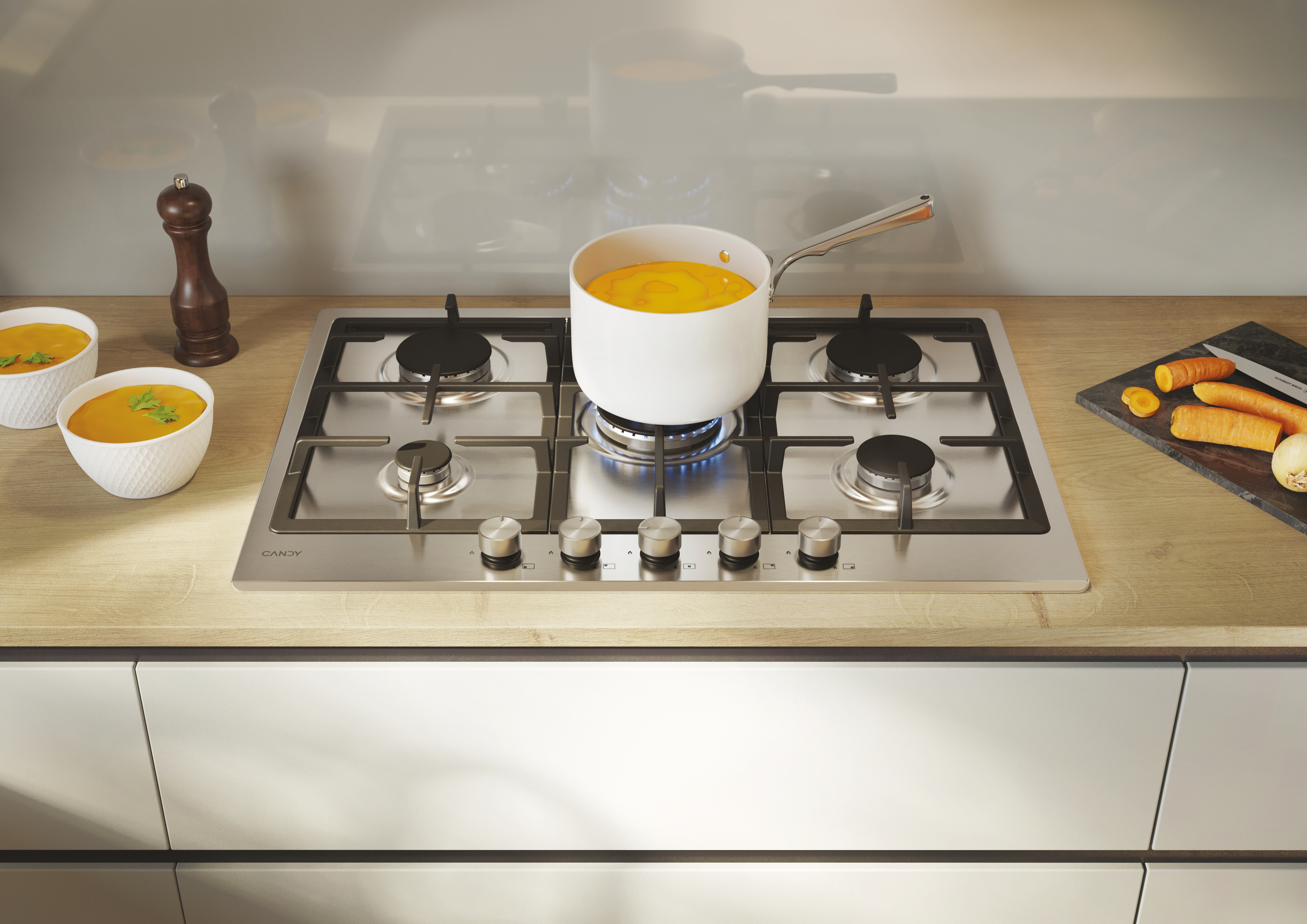 Srebrna płyta grzewcza z pięcioma palnikami. Biały garnek z zupą na palniku. Marchewki i miski obok.