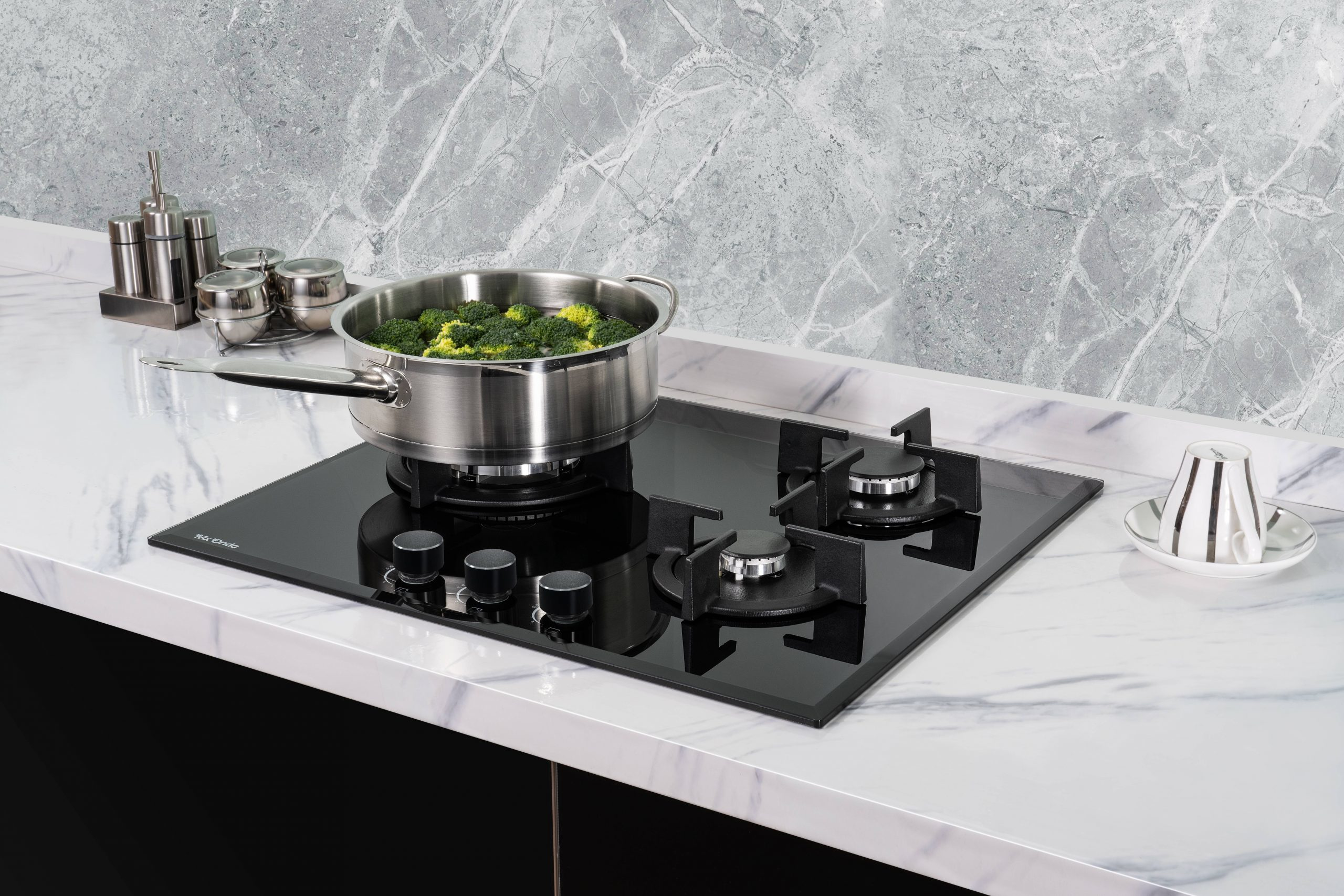 Garnek ze stali nierdzewnej z brokułami na czarnej płycie grzewczej. Marmurowy blat i ściana widoczne.