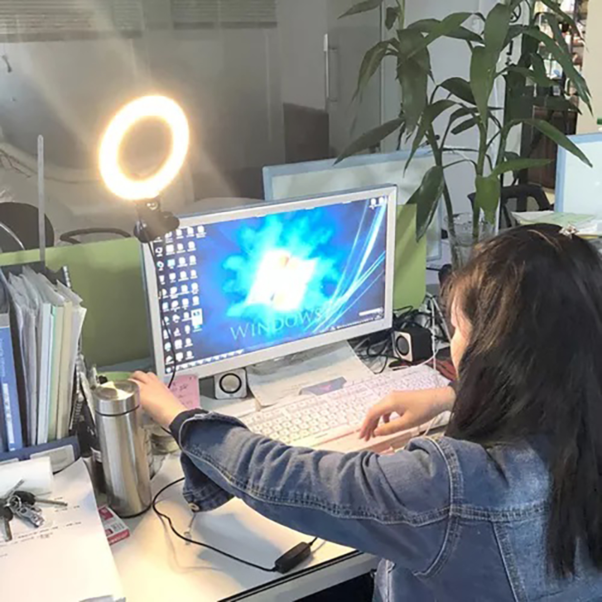 Una mujer está trabajando en una computadora con una luz de anillo. Un termo plateado y un teclado están en el escritorio.