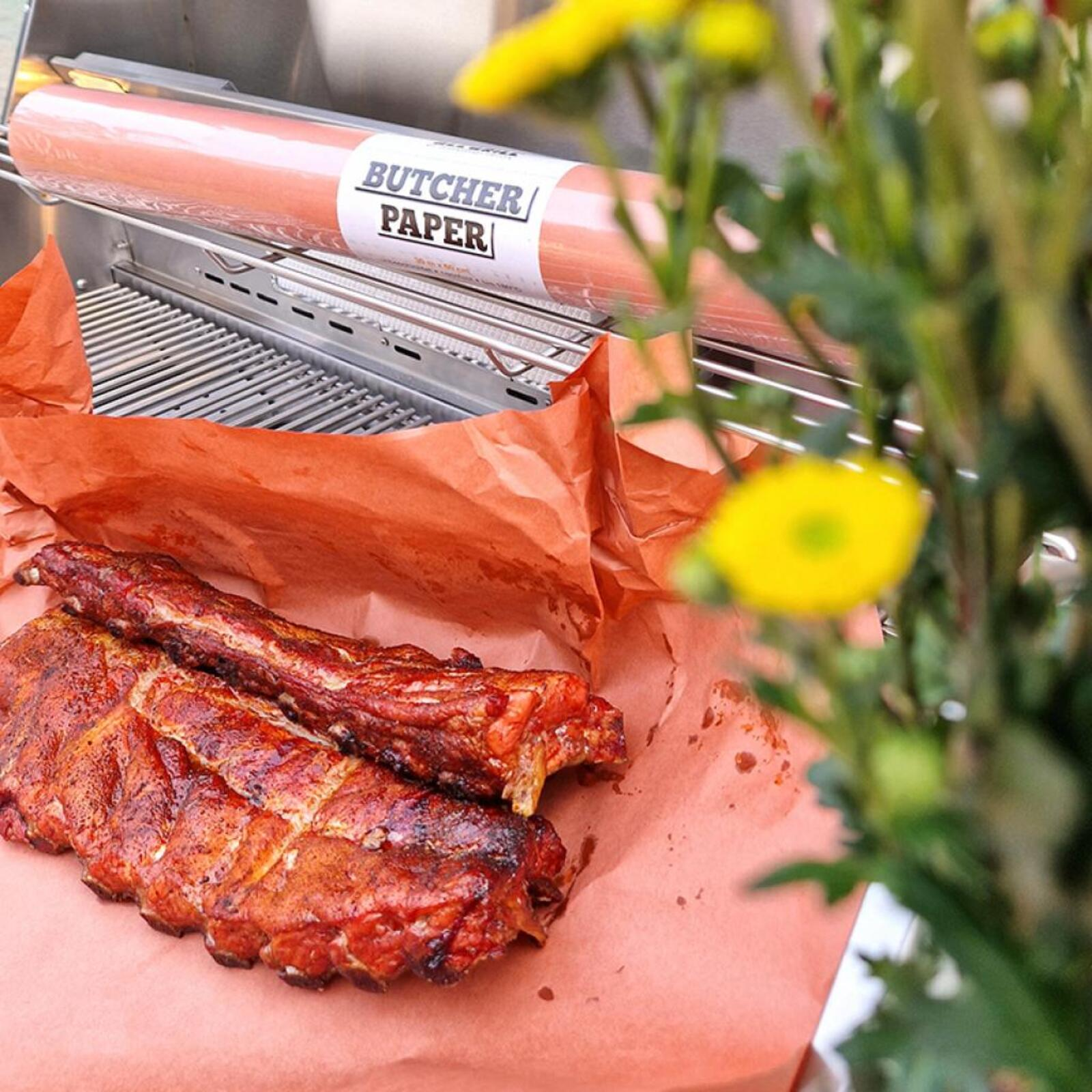 Geräucherte Rippchen auf Metzgerpapier neben einem Grill. Einige gelbe Blumen im Hintergrund.