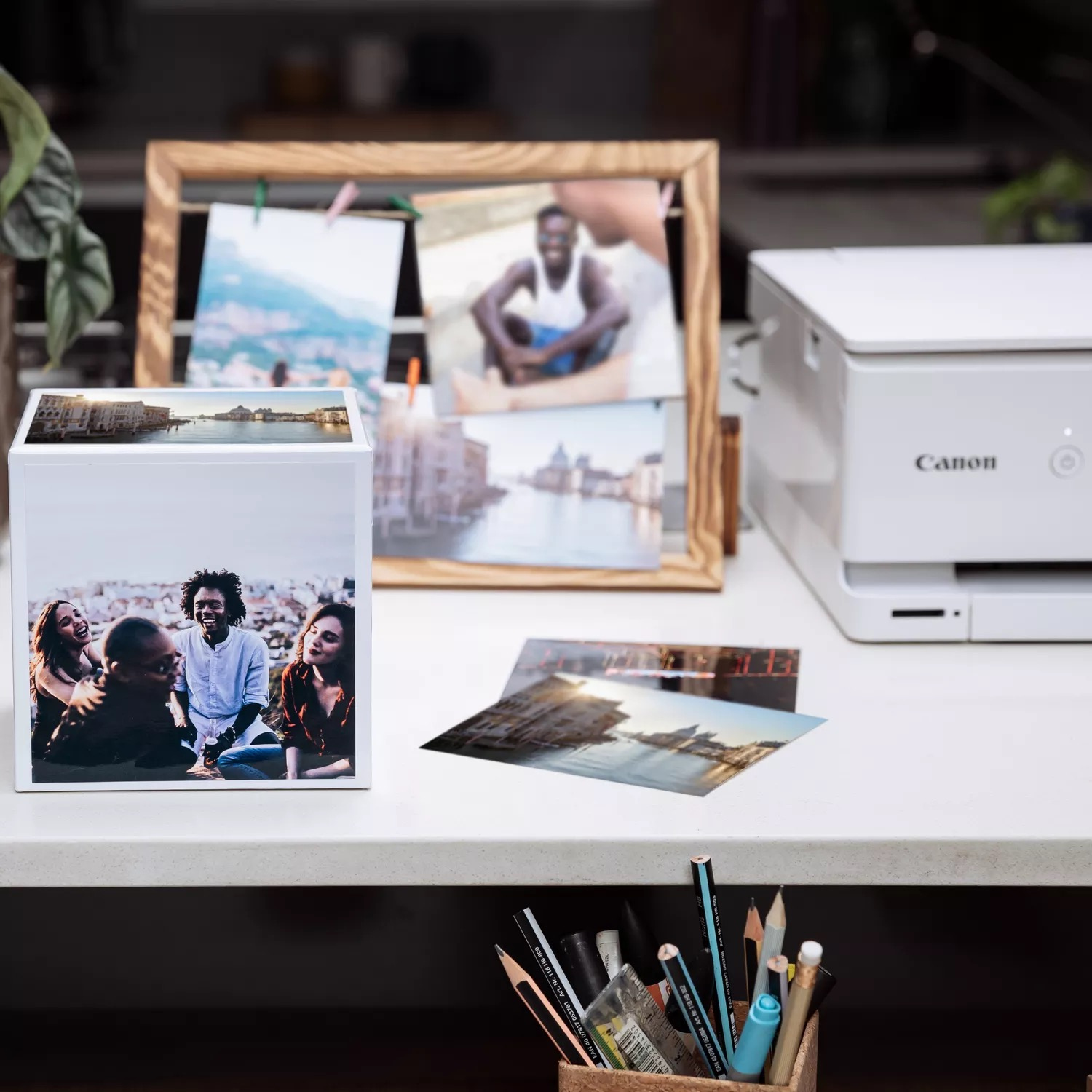 Kostka ze zdjęciami na biurku ze zdjęciami, drukarką, ołówkami i rośliną doniczkową.