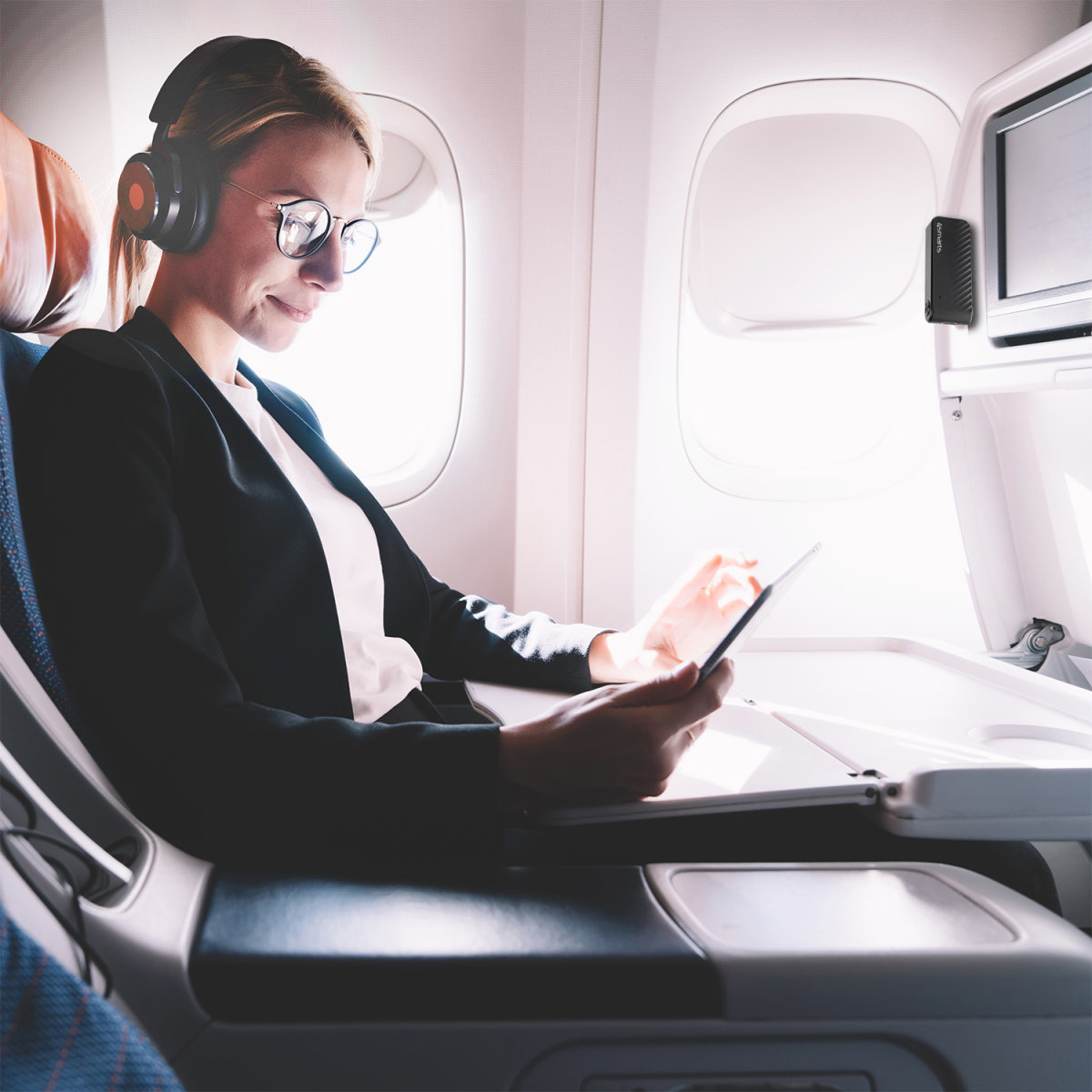 Eine Frau im Flugzeug, mit Kopfhörern, schaut auf ein Tablet in einer gut beleuchteten Kabine.