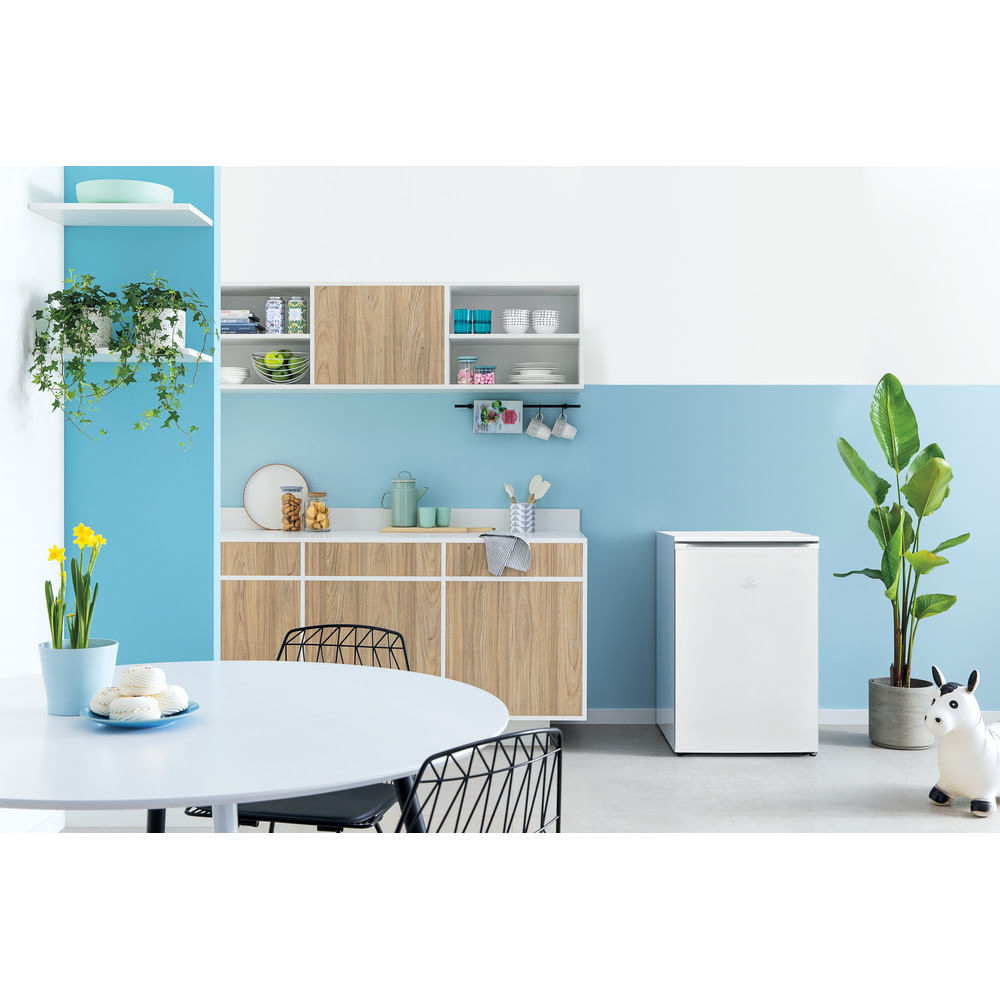 Una cocina con un frigorífico blanco, una mesa redonda, un armario y una planta. Las paredes son azules y blancas.