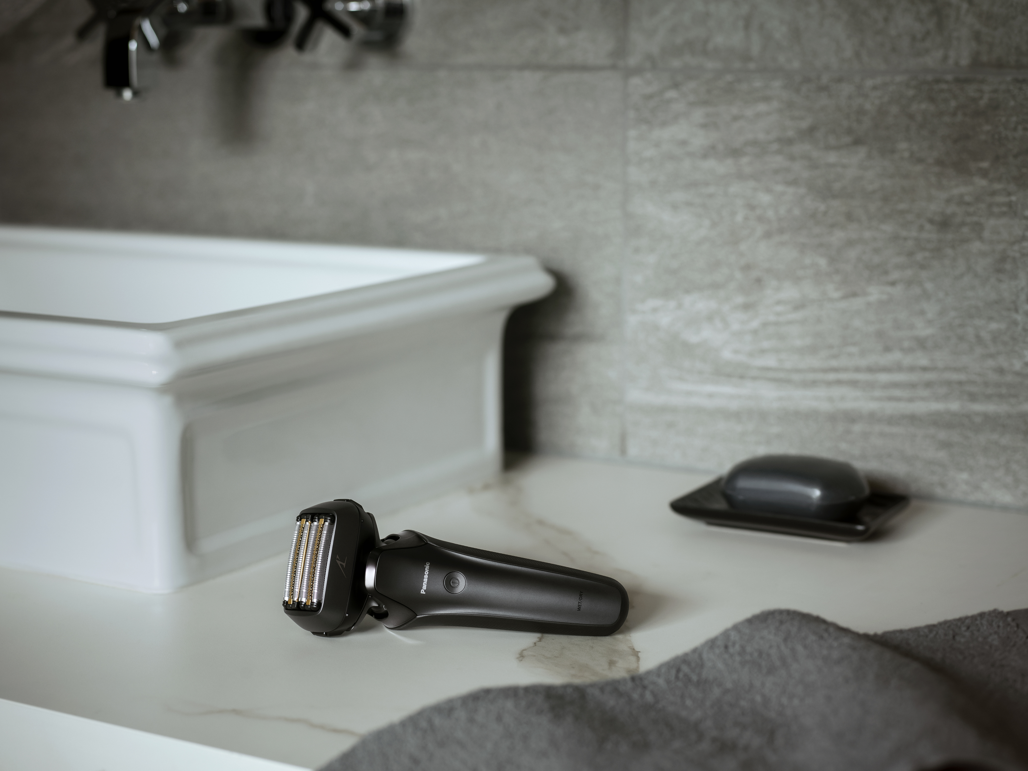 Czarna golarka elektryczna na białym blacie w łazience, z umywalką i mydłem.