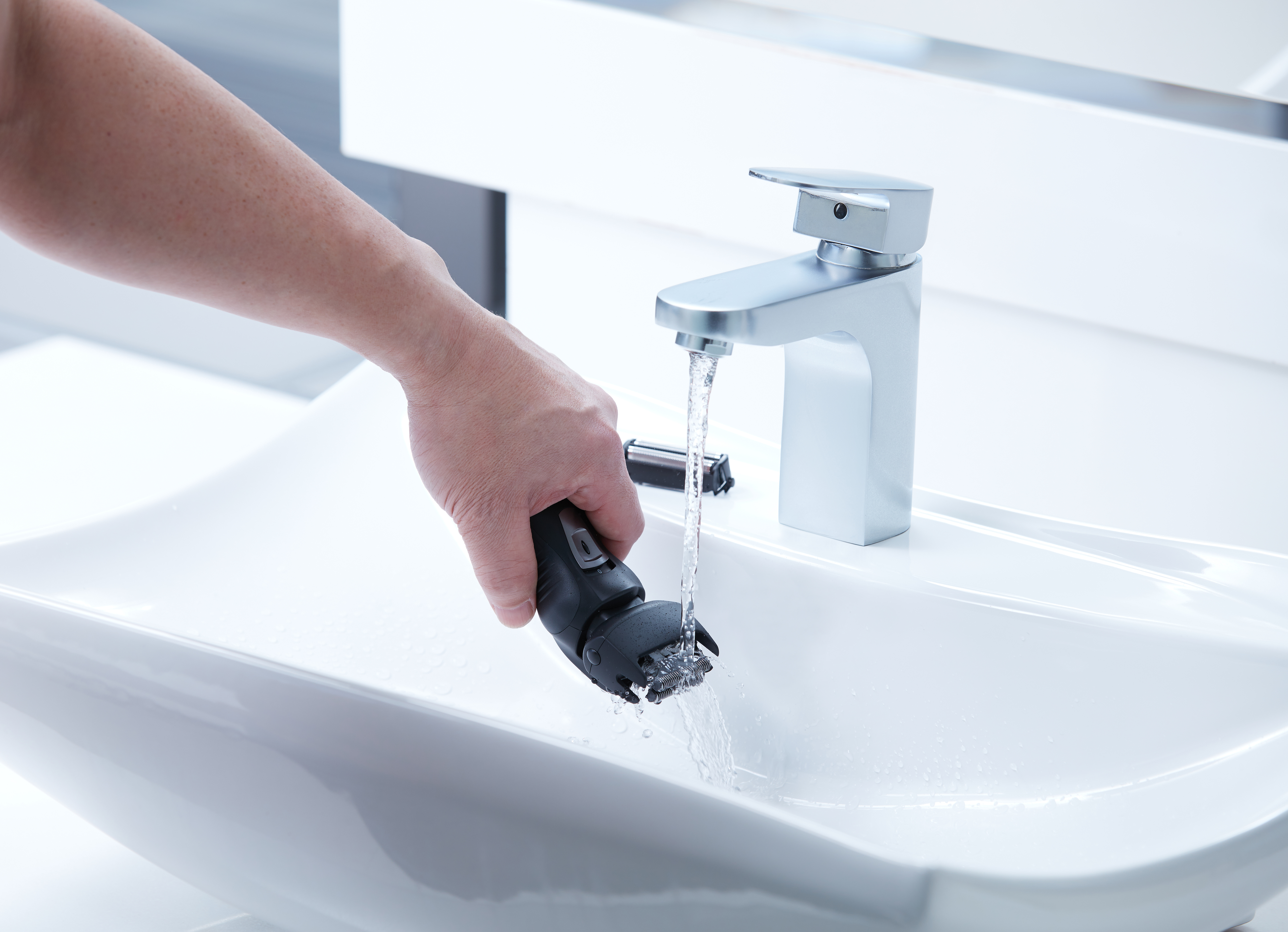 Ręka myjąca czarno-srebrną golarkę elektryczną pod bieżącą wodą w białym zlewie, ze srebrną baterią.