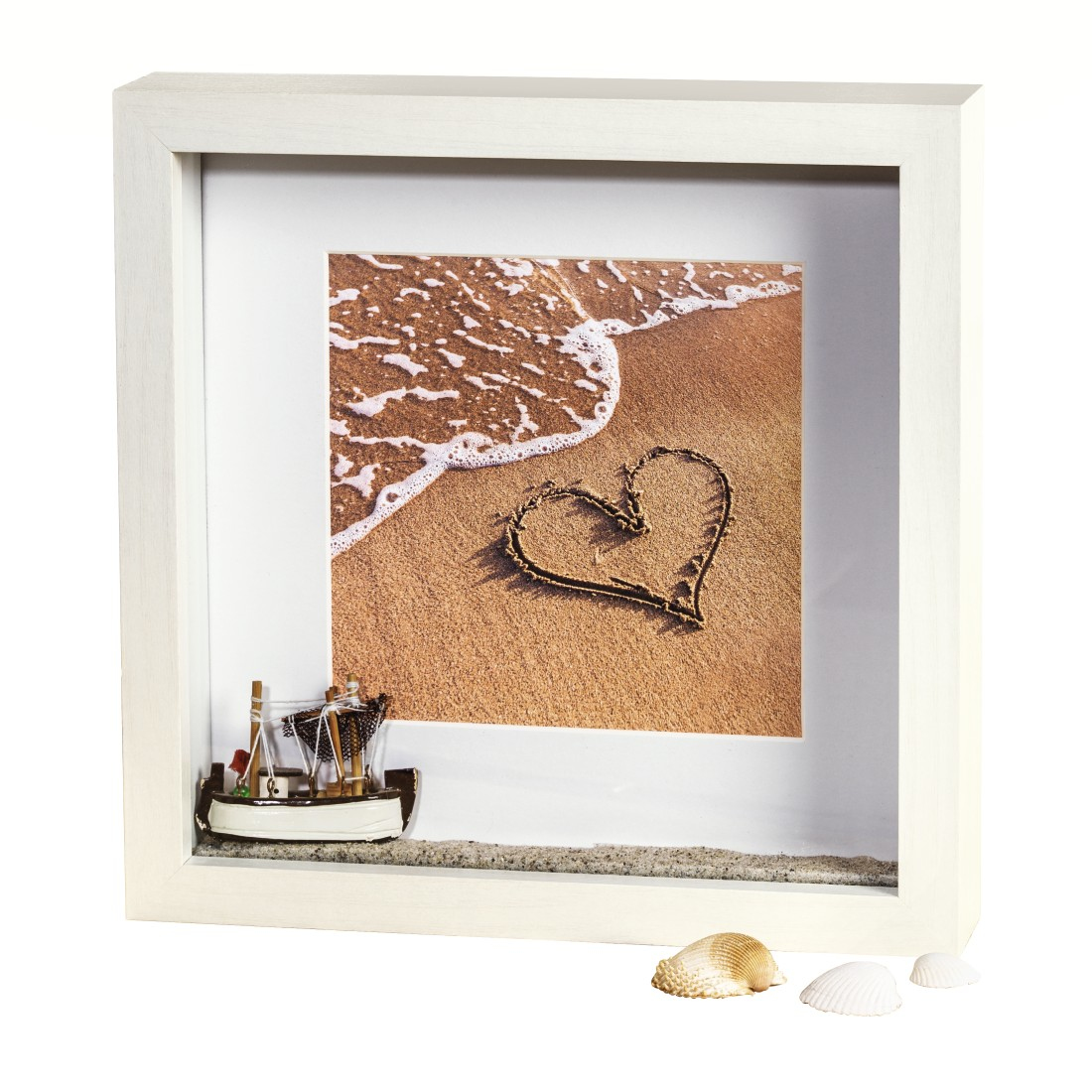 Gerahmtes Bild mit einer Strandszene. Ein Herz ist im Sand gezeichnet. Ein Spielzeugboot und Muscheln davor.