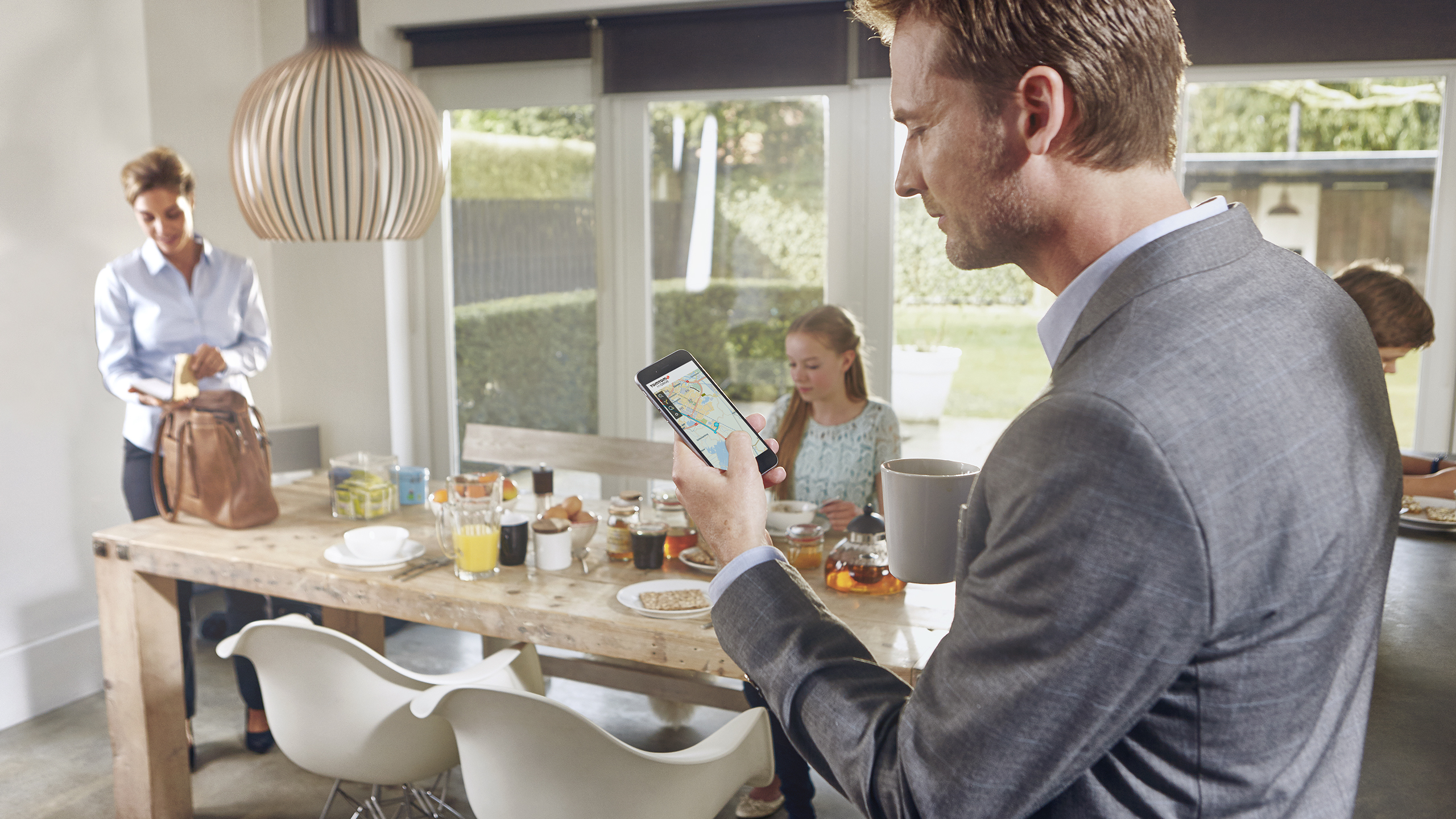 Mann im Anzug hält Telefon. Andere frühstücken. Auf dem Telefon ist TomTom-Gerät.