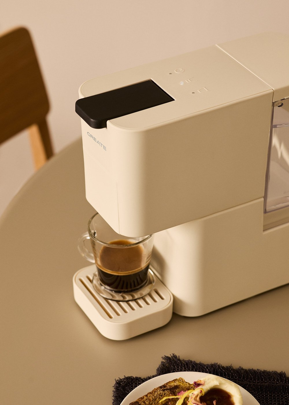 Cafetera crema con la palabra CREATE, y un vaso de café. Mesa beige, servilleta negra.