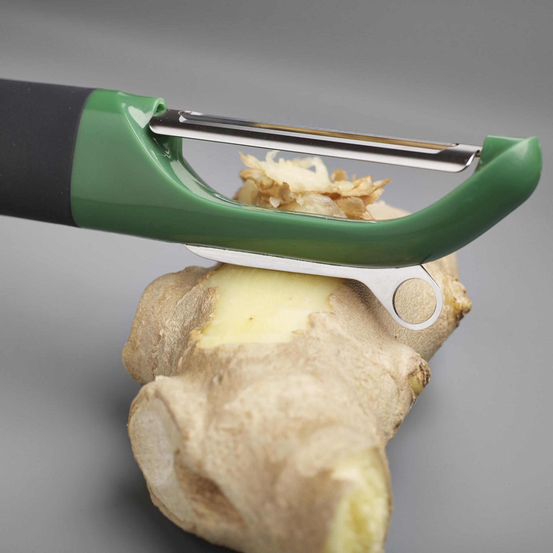 A green and black peeler is peeling a piece of ginger on a gray background.