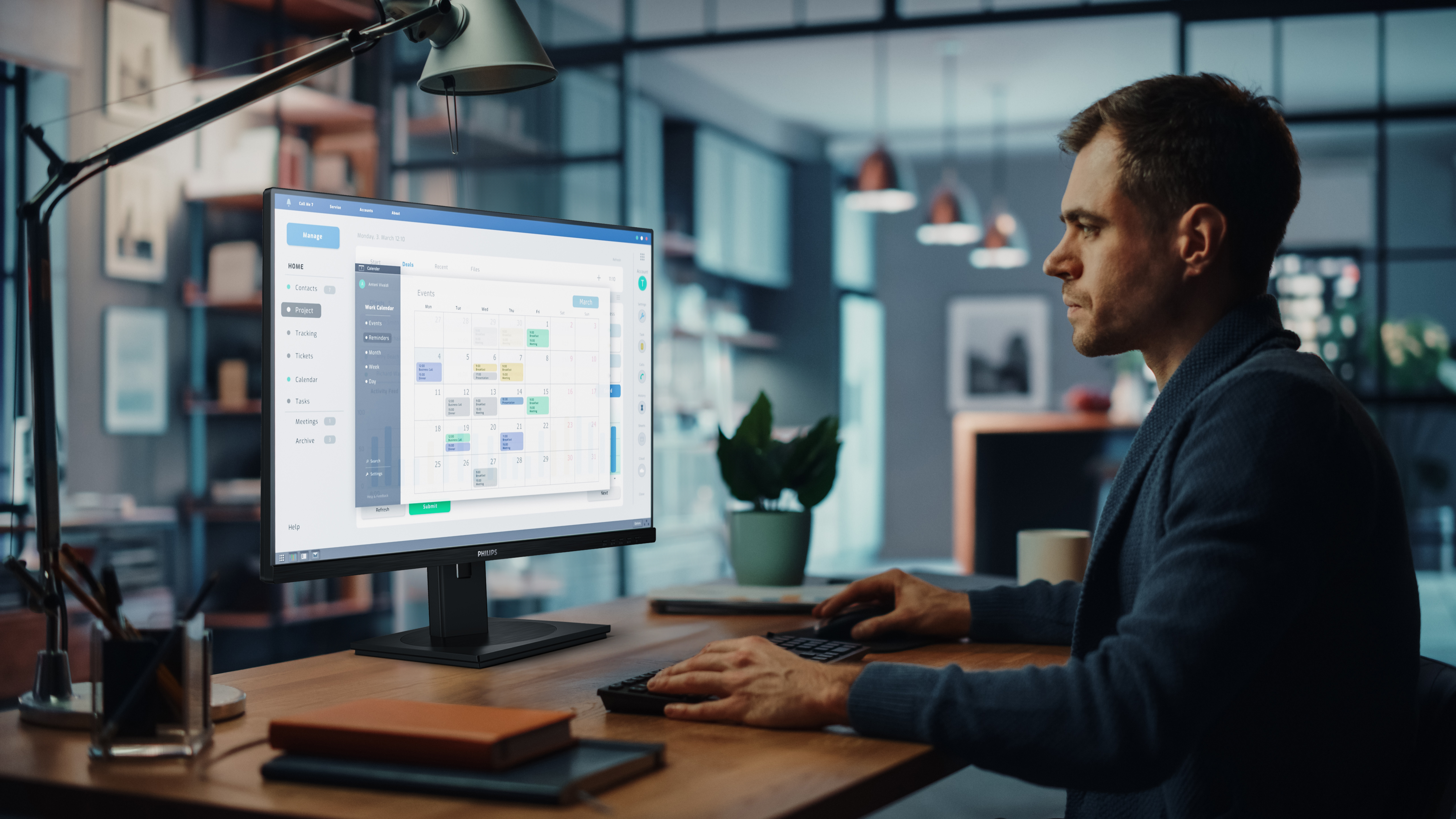 Mężczyzna siedzi przy biurku i pracuje na monitorze wyświetlającym kalendarz. Na biurku znajduje się lampa i filiżanka.